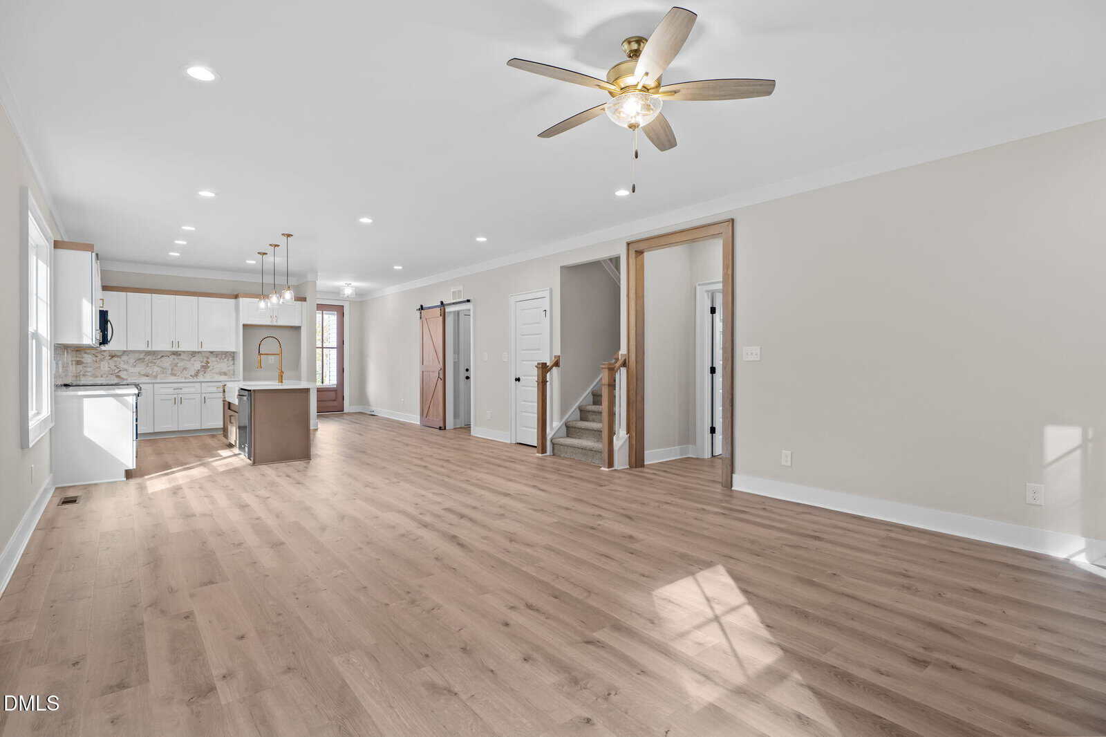 Open-concept living space with light wood floors, white walls, and ceiling fan. Modern kitchen with white cabinets and island, farmhouse-style sink, and pendant lights. Staircase and sliding barn door in the background.