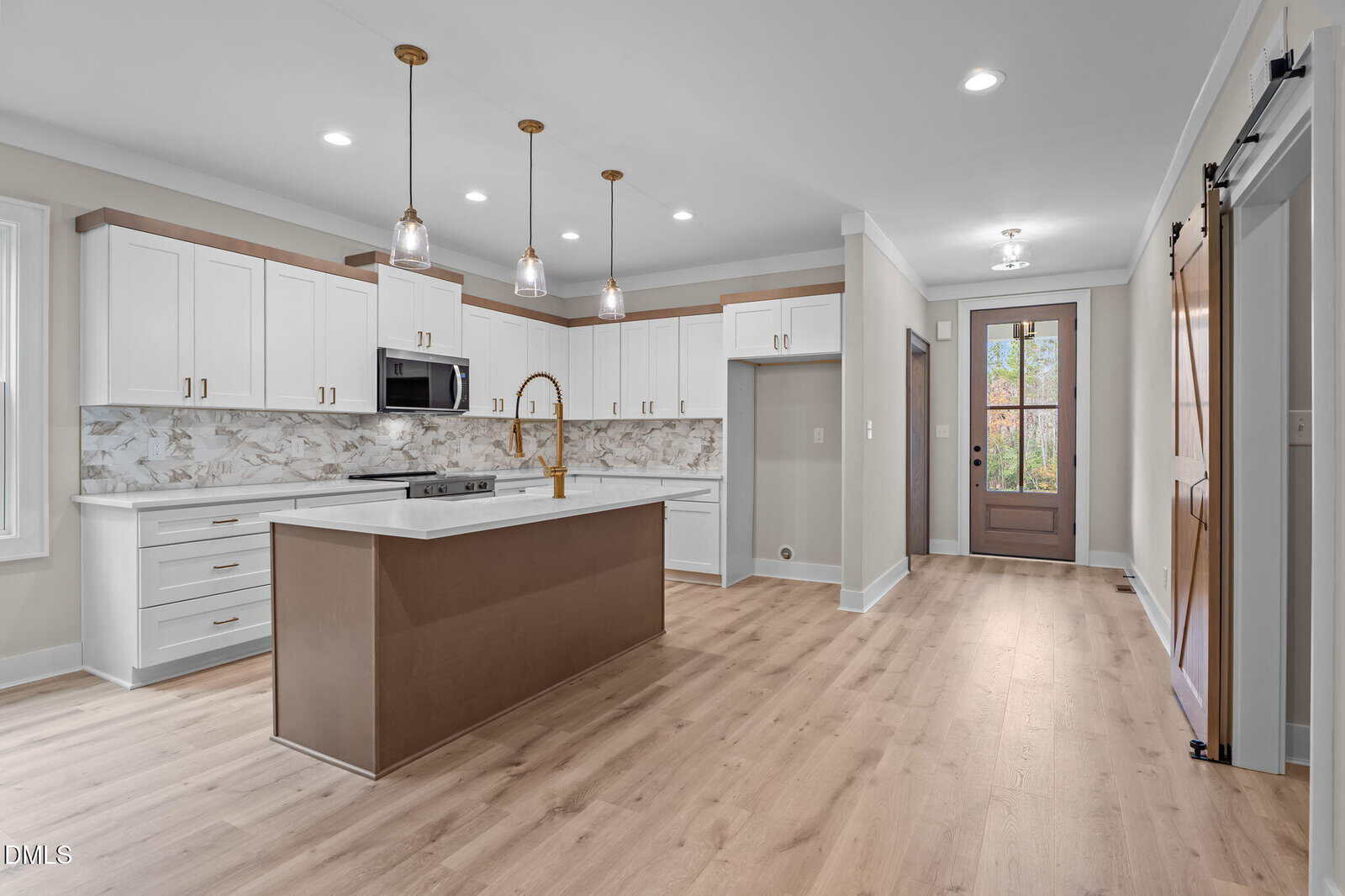 Modern kitchen with white cabinets, marble backsplash, center island with brown base, gold faucet, pendant lights, light wood flooring, and a wooden sliding barn door near a glass front entry door.