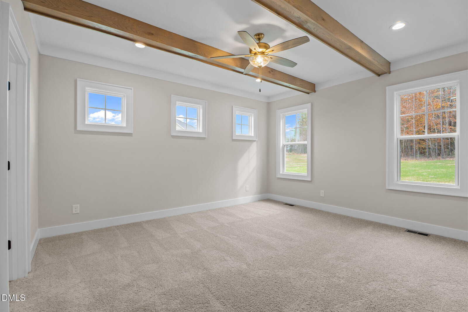 Bright, empty room with beige carpet, white walls, three small square windows, one large window, ceiling fan with lights, and exposed wooden ceiling beams. Trees and grass visible outside.