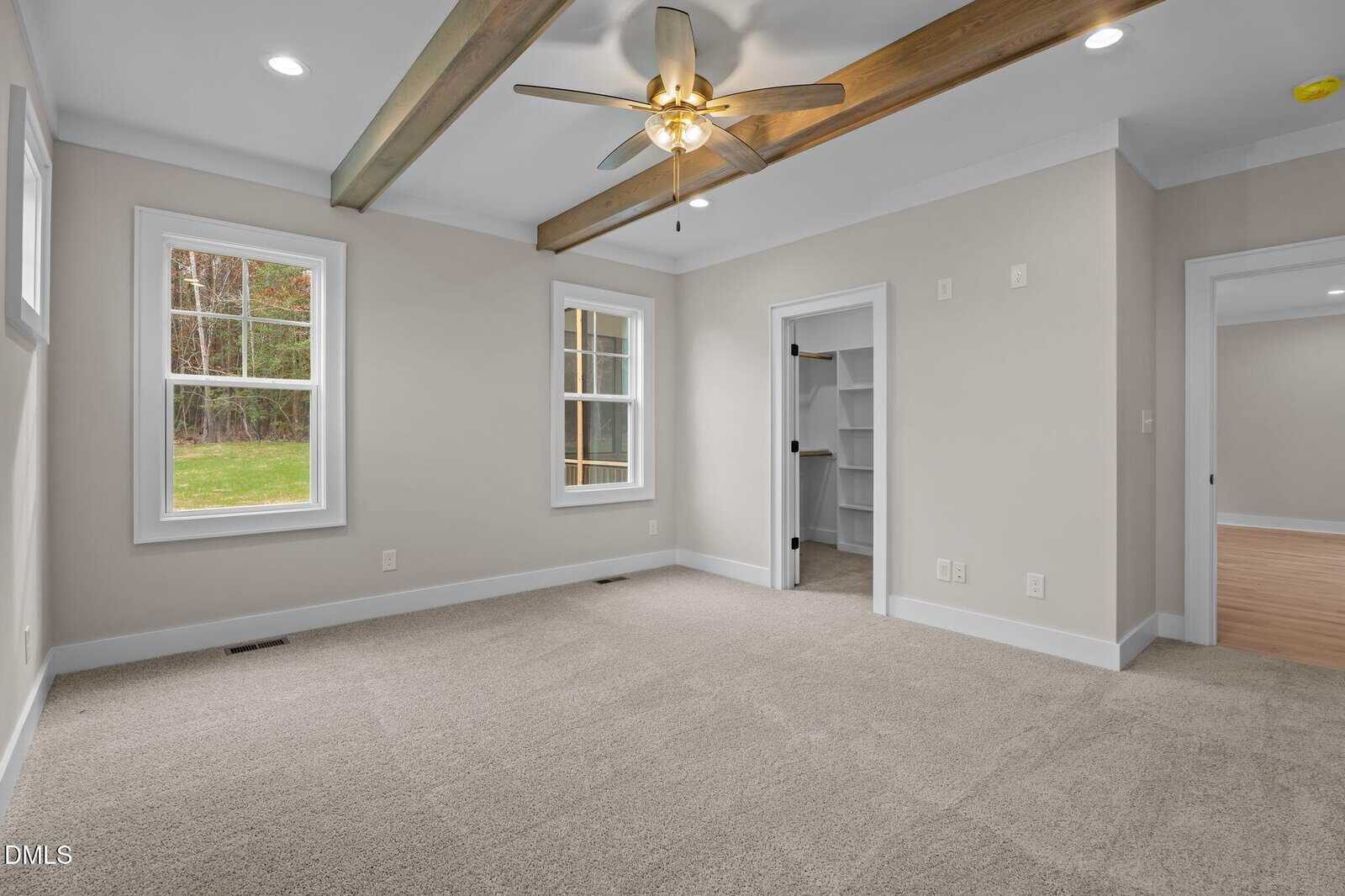 Bright, empty room with beige carpet, white walls, wood ceiling beams, a ceiling fan, two windows, and an open door leading to a walk-in closet with shelves. Another open doorway leads to a hallway with wood flooring.