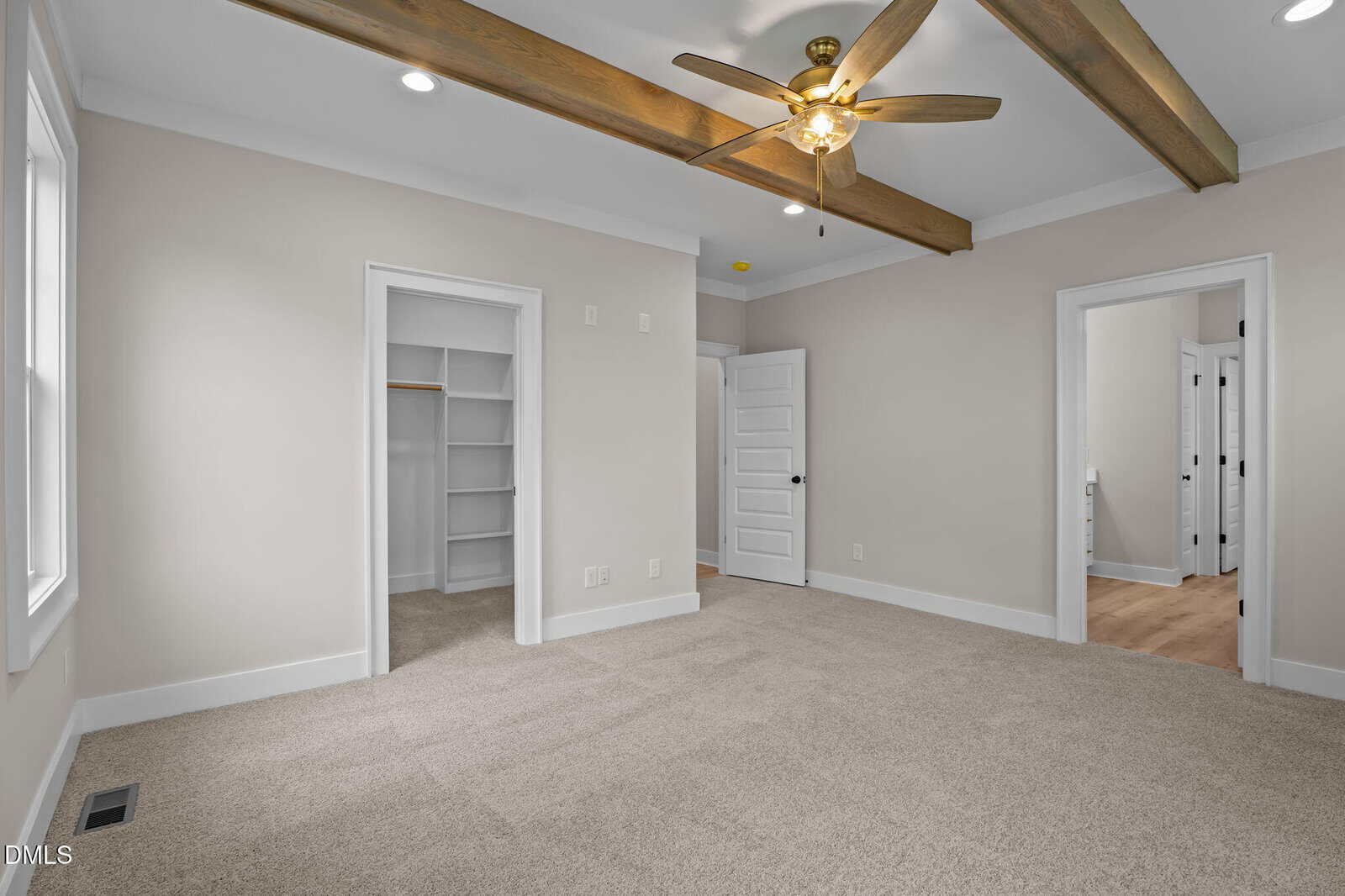 A spacious, carpeted bedroom with beige walls, wood ceiling beams, a ceiling fan, open closet, and two doorways leading to other rooms. Three windows provide natural light.