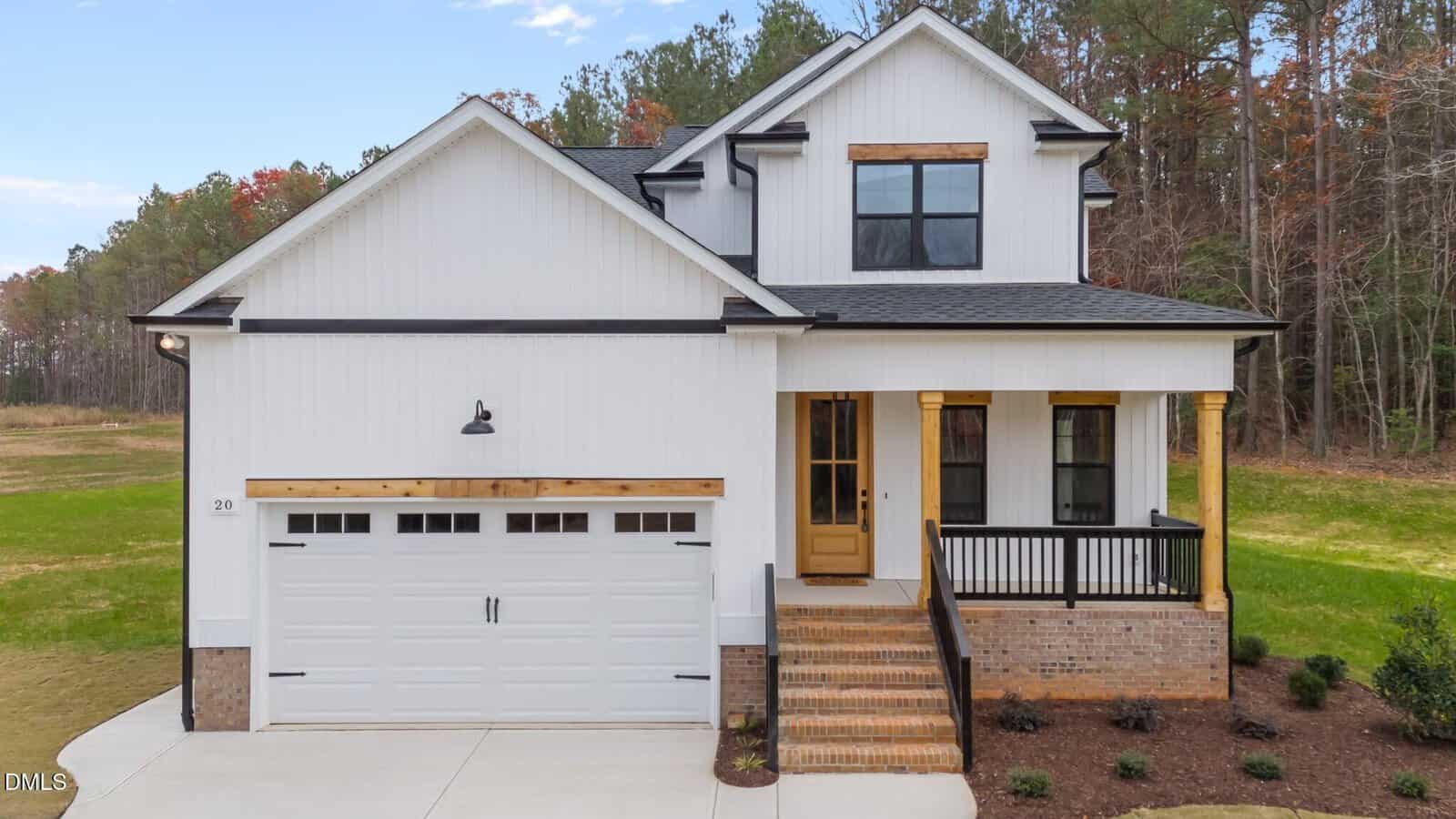 A modern white two-story house with black trim, wood accents, a front porch, double garage, brick steps, and a landscaped yard, set against a backdrop of green trees and lawn.