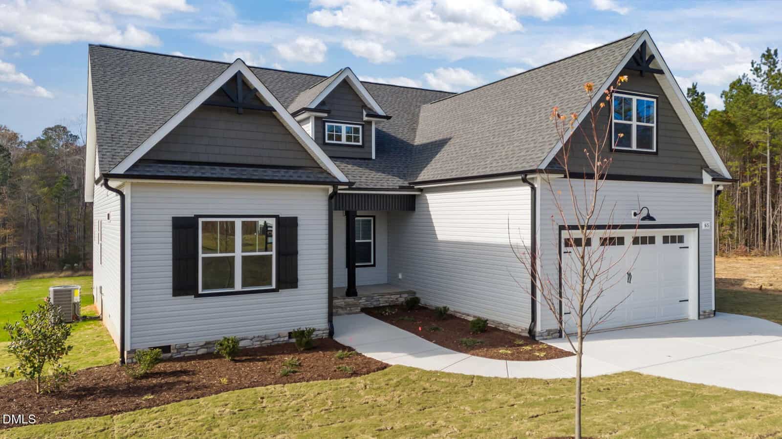 A modern suburban house with gray siding, black shutters, a two-car garage, and a gabled roof, surrounded by a grassy yard and young landscaping under a partly cloudy sky.