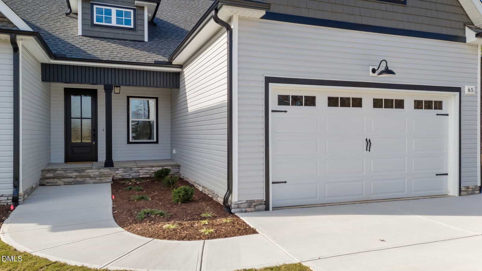 A modern home exterior featuring a two-car white garage door with windows, light gray siding, stone accents, black trim, a black front door, and a small landscaped area next to the entrance.