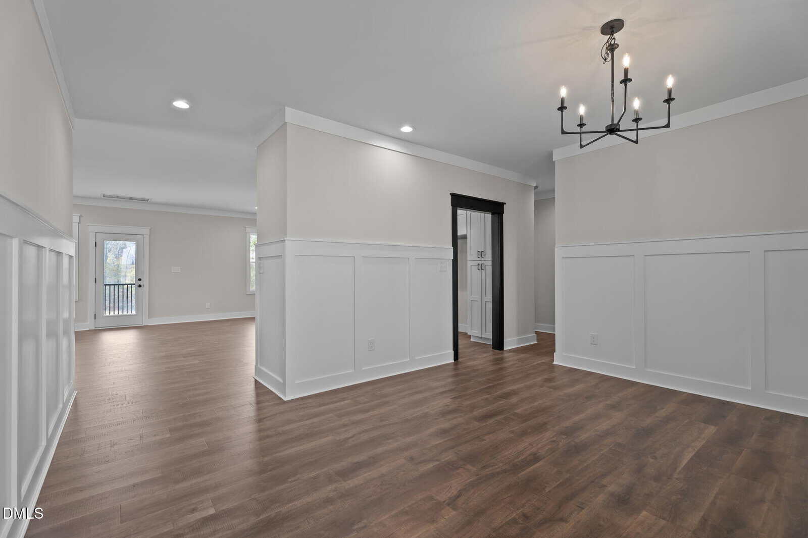 A modern open-concept room with light gray walls, white wainscoting, wood flooring, a black-framed doorway, and a black chandelier. Natural light enters from windows in the adjacent space.