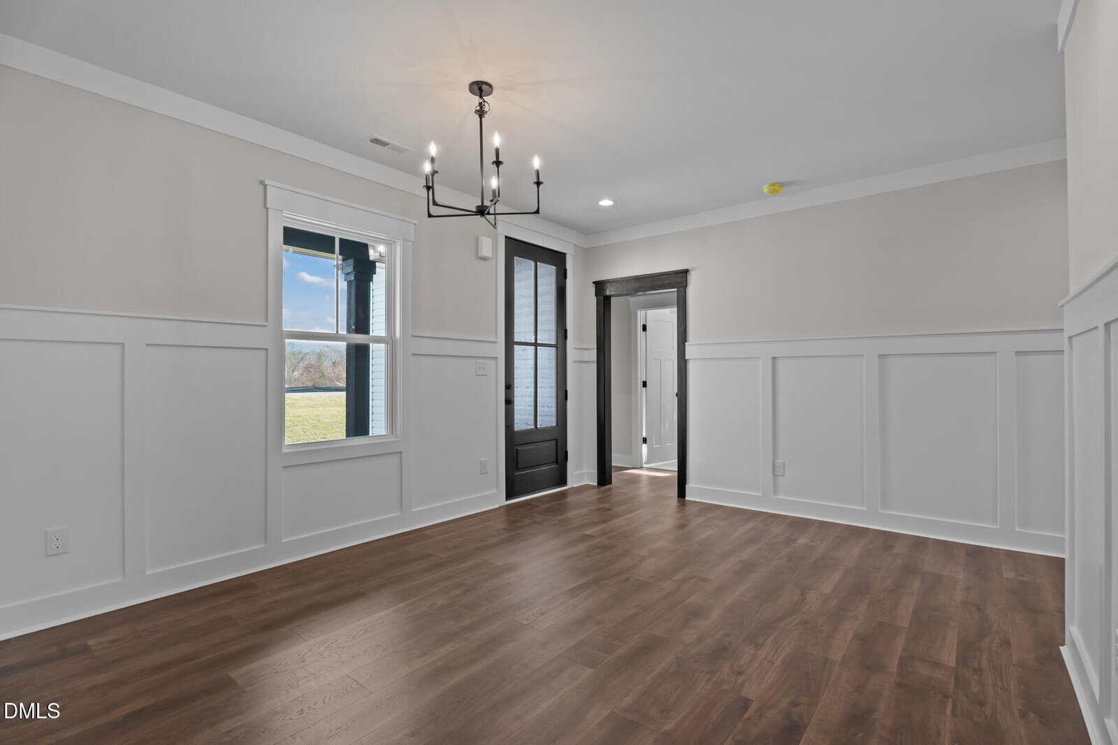 A bright, empty room with wood floors, white wainscoting, beige walls, a black chandelier, a large window, and a black door leading to a hallway with more doorways.