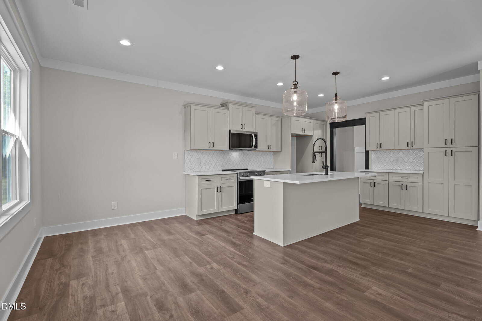 Modern kitchen with light gray cabinets, white island and countertops, dark appliances, pendant lights over the island, and wood-look flooring. Large window on the left lets in natural light.