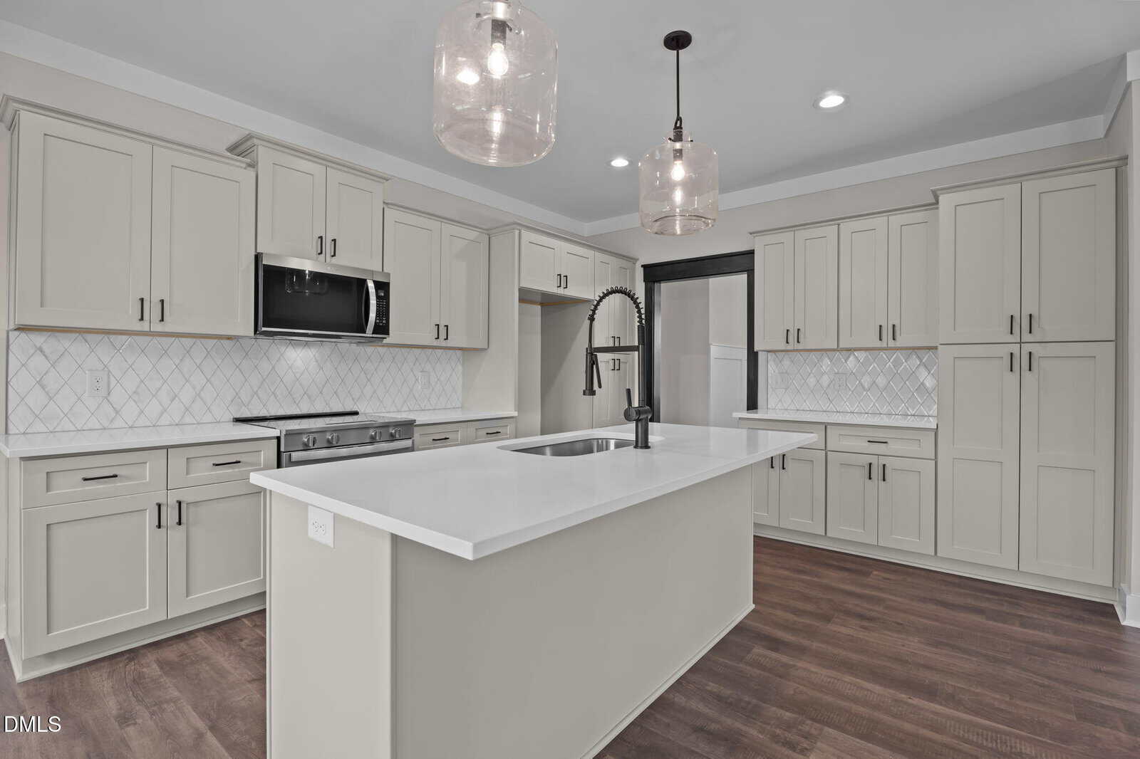 Modern kitchen with light gray cabinets, stainless steel appliances, white countertops, a large island with a sink and black faucet, pendant lights, white tile backsplash, and dark wood flooring.