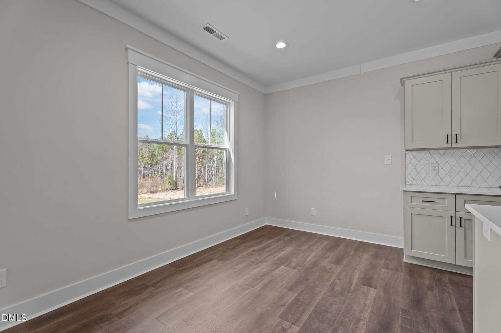 A bright room with large window, light gray walls, dark wood floors, and a partial view of a kitchen with white cabinets and a tiled backsplash. Trees and a blue sky are visible outside the window.
