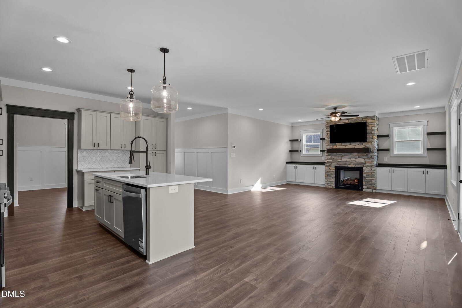 Open-concept living area with wood floors, a modern kitchen island, pendant lights, and a stone fireplace beneath a wall-mounted TV. White cabinets and natural light enhance the spacious, airy feel.