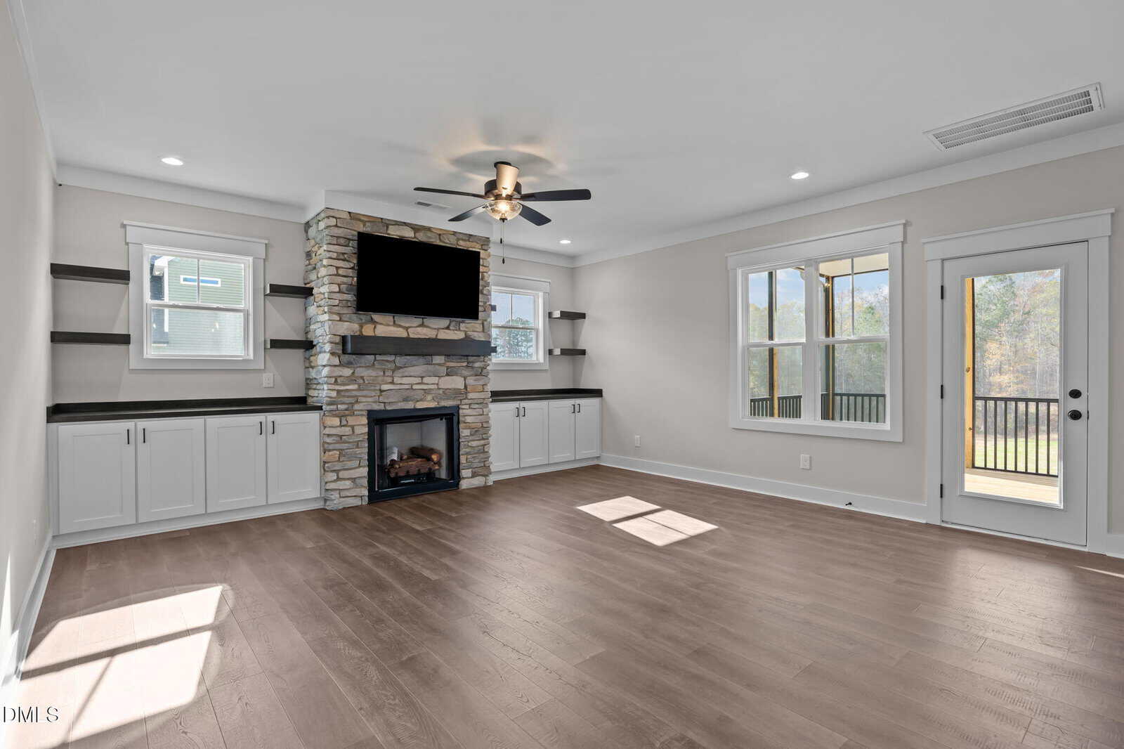 A bright living room with wood floors, a ceiling fan, stone fireplace, mounted TV, built-in shelves and cabinets, large windows, and a glass door leading to a deck with a view of trees outside.