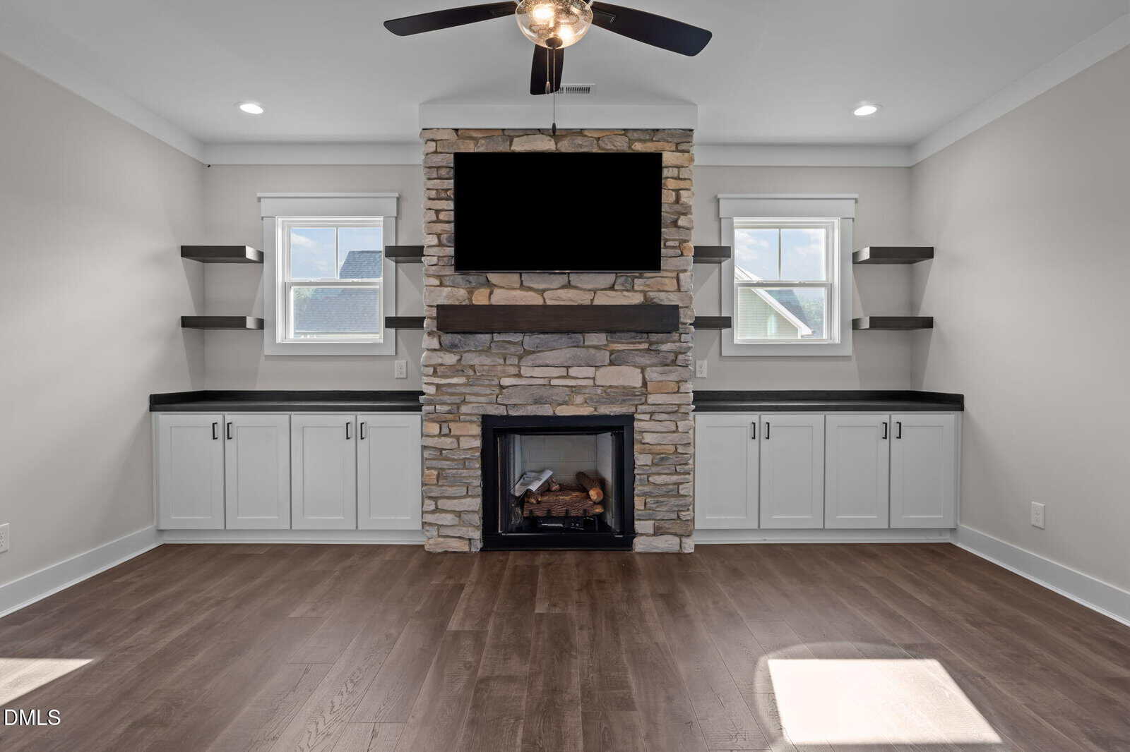Modern living room with stone fireplace, wall-mounted TV above, built-in cabinets with black countertops, floating shelves, two windows, ceiling fan, and wood flooring.
