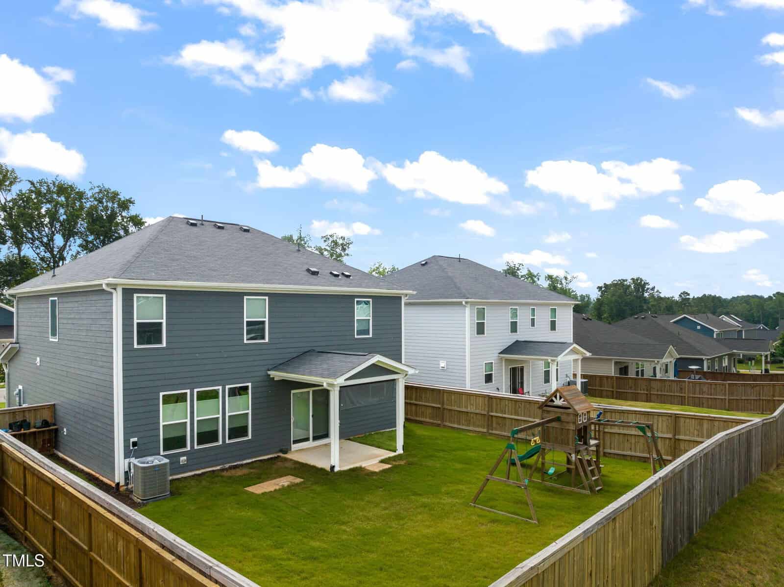 A fenced backyard with green grass, a wooden playset with swings, and a slide. Behind the yard is a two-story gray house with covered patio and white trim. Other houses and trees are visible in the background under a blue sky.