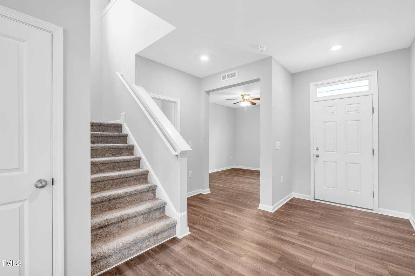 Bright entryway with wood flooring, white walls, and recessed lighting. Carpeted stairs lead up to the left, and a doorway reveals a ceiling fan in an adjacent room. A closed white front door is to the right.