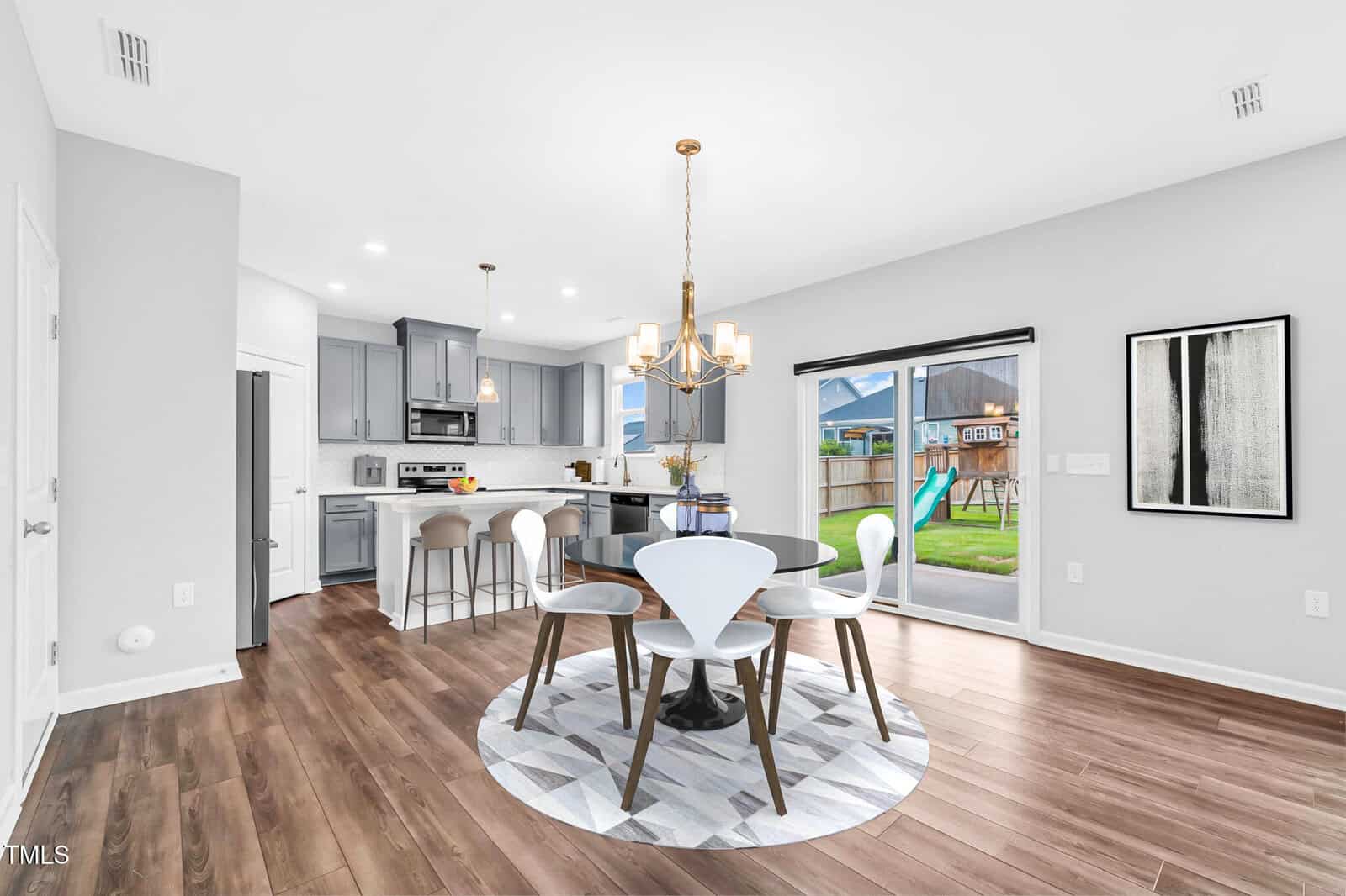 Modern open kitchen and dining area with wood floors, gray cabinets, round dining table, white chairs, contemporary chandelier, large window with backyard view, and abstract artwork on the wall.