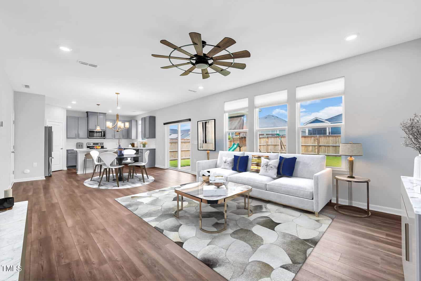 Modern open-concept living and dining area with wood flooring, gray and white decor, large windows letting in natural light, and a view of a fenced backyard. A ceiling fan hangs in the center of the space.