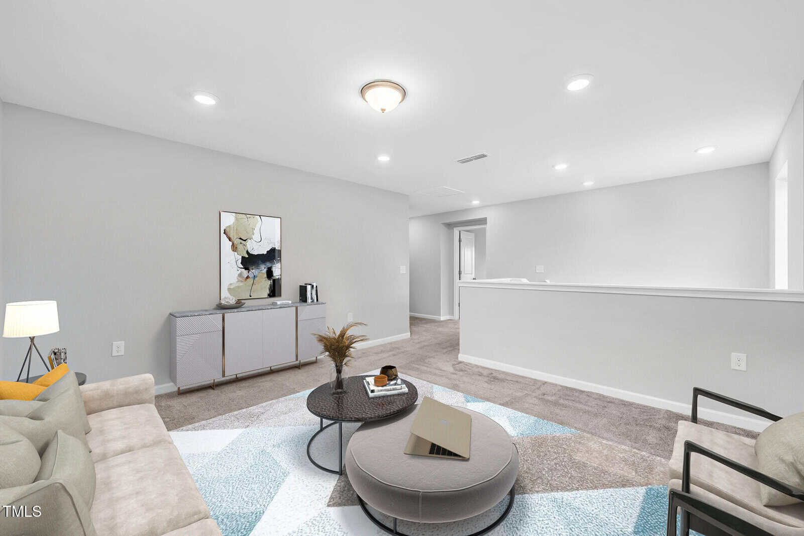 A modern living room with light gray walls, beige carpet, a beige sofa, round ottoman coffee table, two chairs, a sideboard with abstract art, and recessed ceiling lights. The space is bright and minimally decorated.
