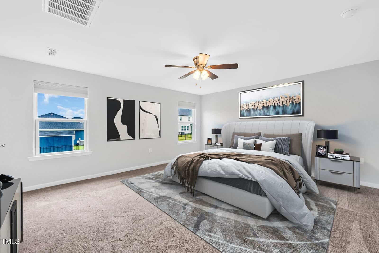 A modern, spacious bedroom with a large bed, gray upholstered headboard, abstract wall art, two windows with outside views, and a ceiling fan with a light. Room features neutral colors and soft carpet flooring.