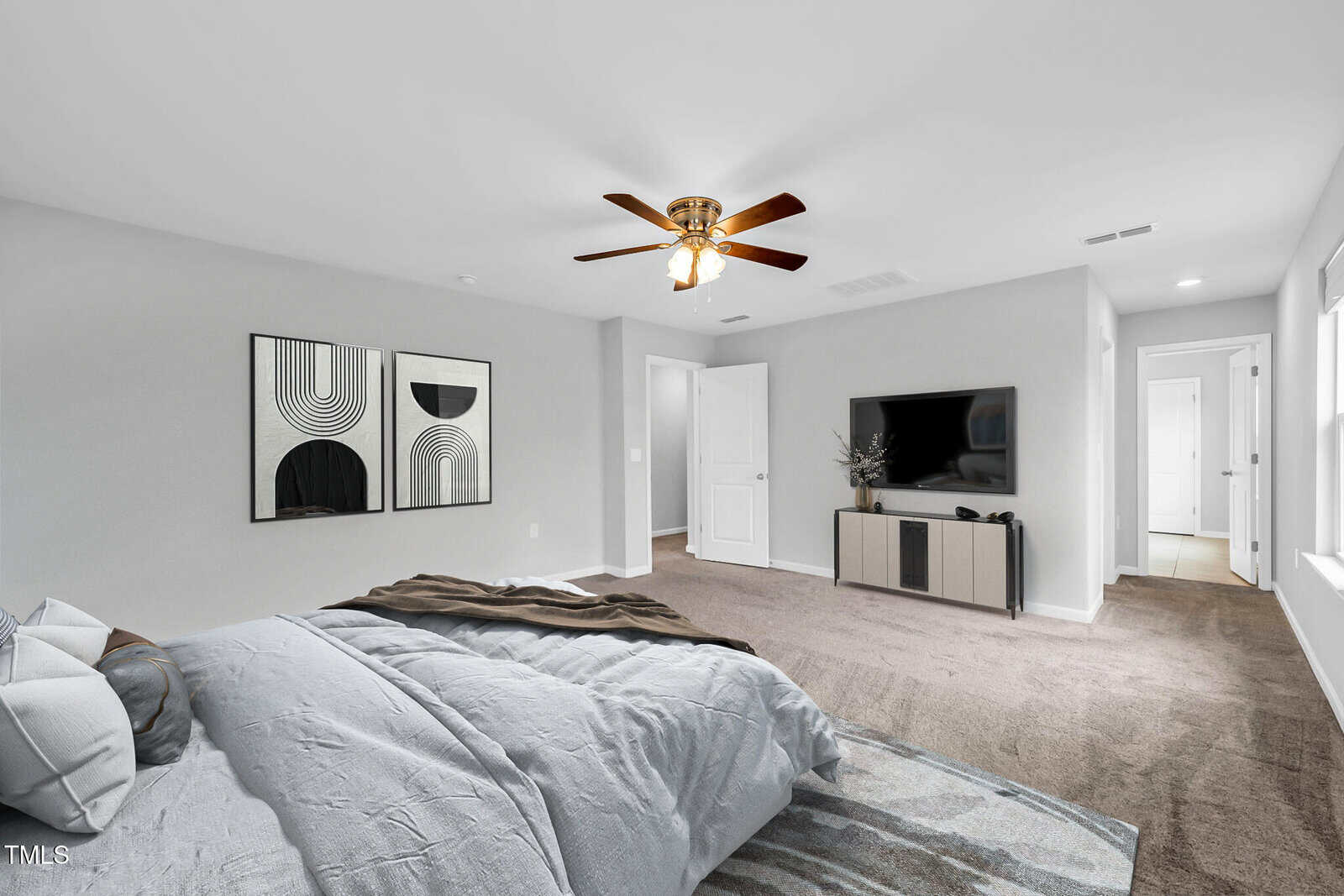 A spacious, modern bedroom with a large bed, abstract art on the wall, a ceiling fan with lights, a TV on a console, and windows letting in natural light. The room has neutral tones and plush carpet.