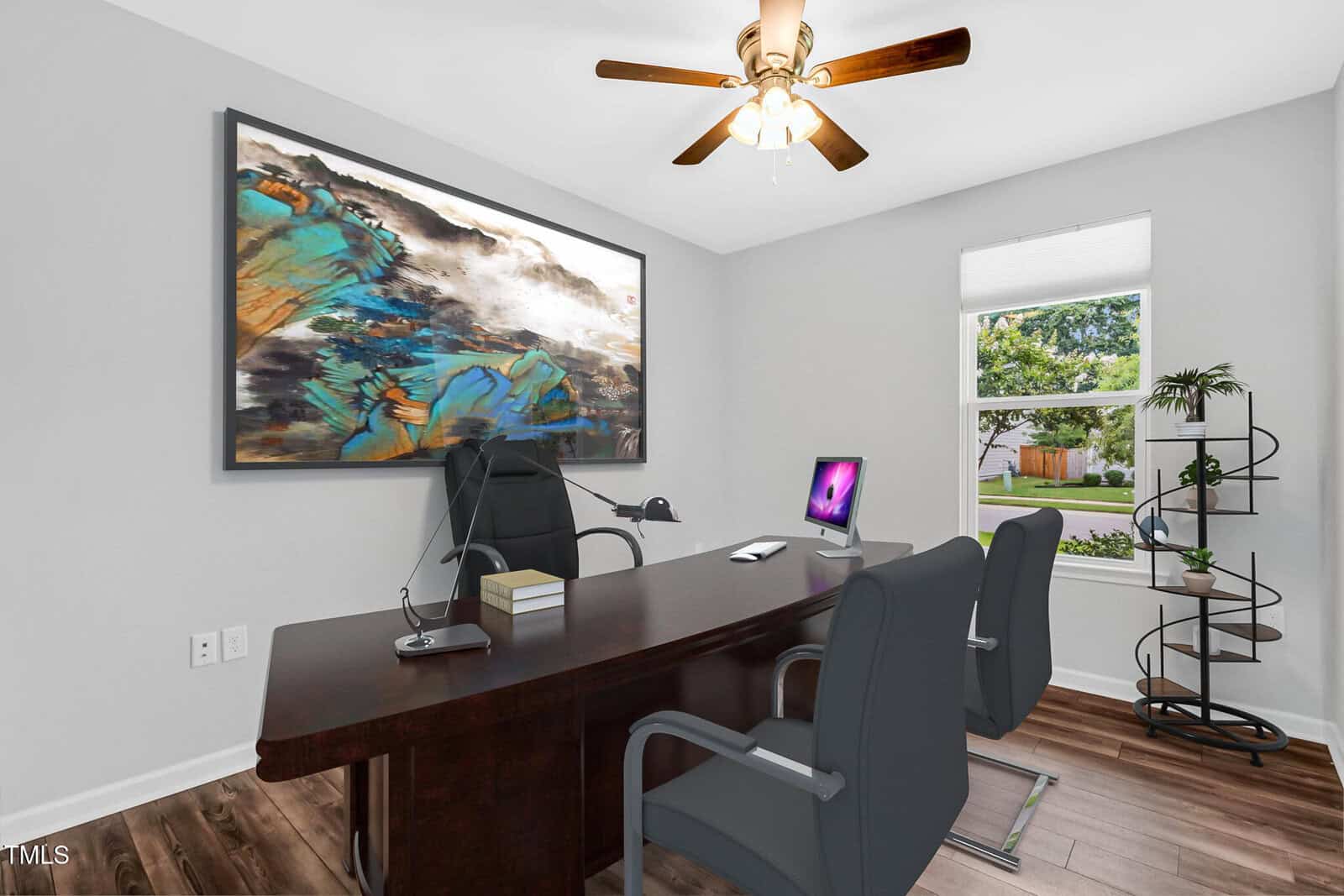 A modern office with a large wooden desk, two gray chairs, a computer, books, and a phone. There is abstract art on the wall, a ceiling fan above, and a window with a view of greenery. A curved plant stand is in the corner.