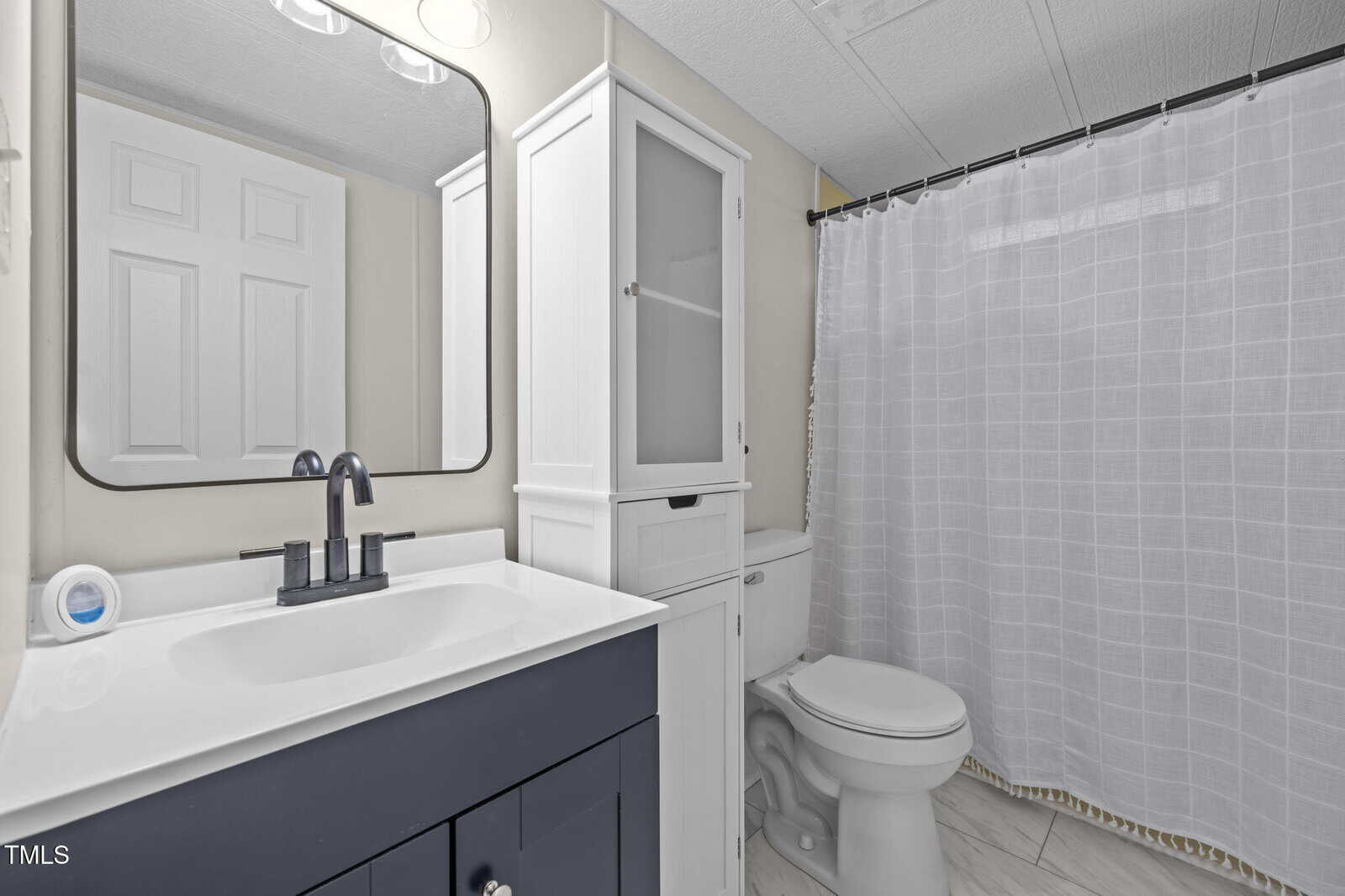 A bathroom with a dark vanity and white countertop, a rectangular mirror, a white cabinet over the toilet, a closed shower curtain, and light-colored tile flooring.