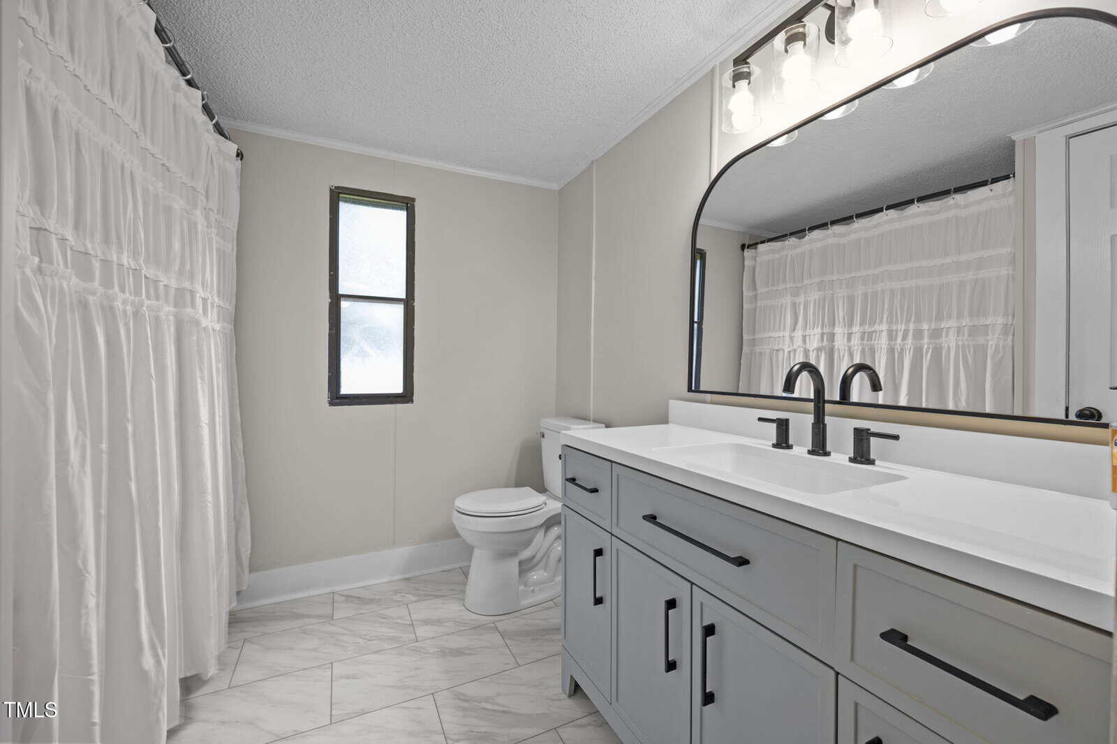 A modern bathroom with a double-sink vanity, black fixtures, a large mirror, a white toilet, a window, and a bathtub with a white shower curtain. The floor is covered with light-colored tiles.