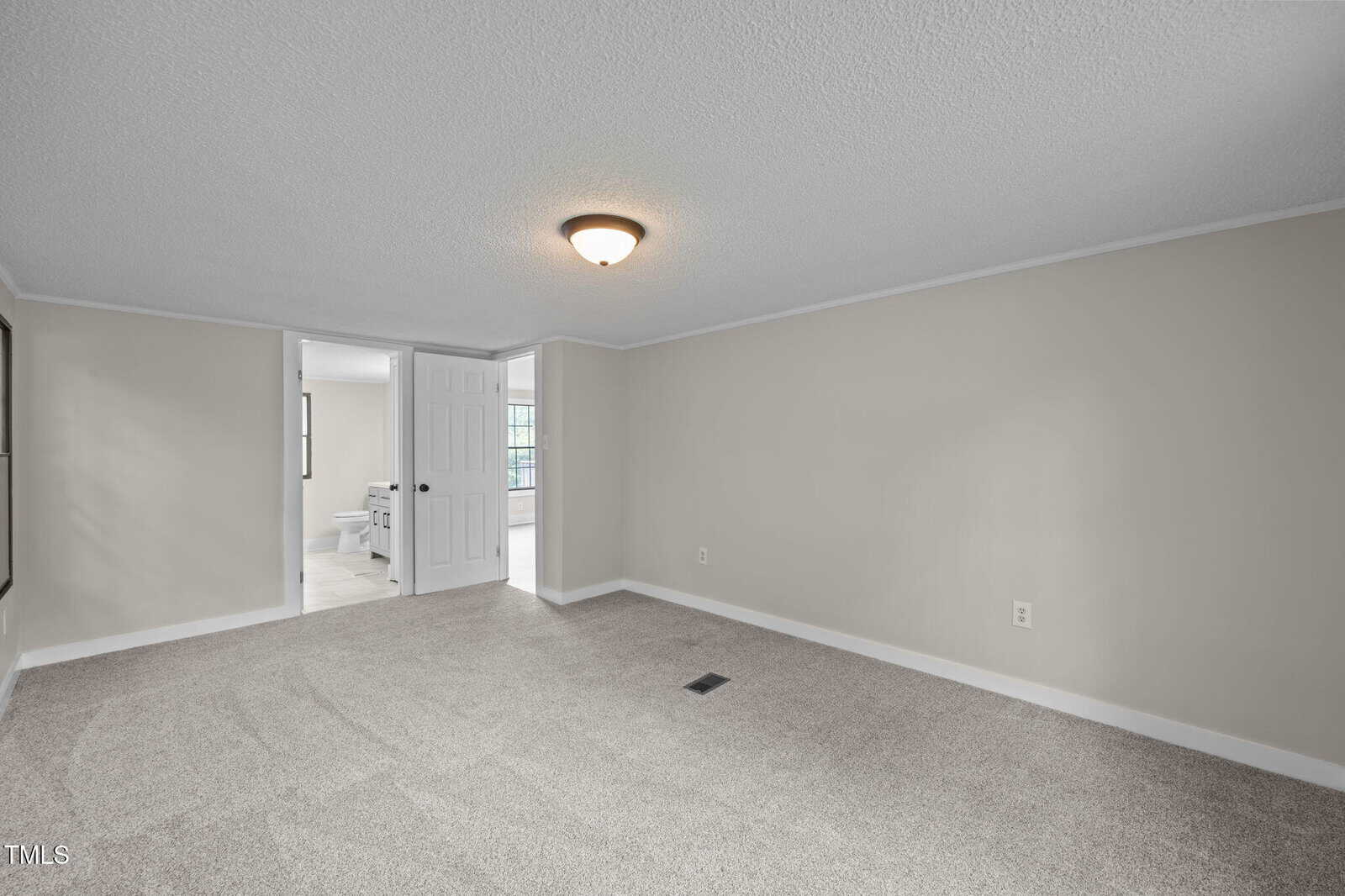 A carpeted, empty room with beige walls, white trim, and a ceiling light fixture. The room has an open doorway leading to a bathroom and another adjacent room with tile flooring visible.