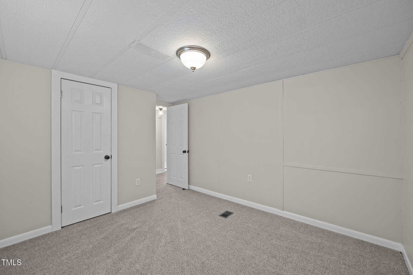 Small, carpeted room with beige walls, white trim, two closed doors, a ceiling light fixture, and a vent on the floor. The room is empty and well-lit.