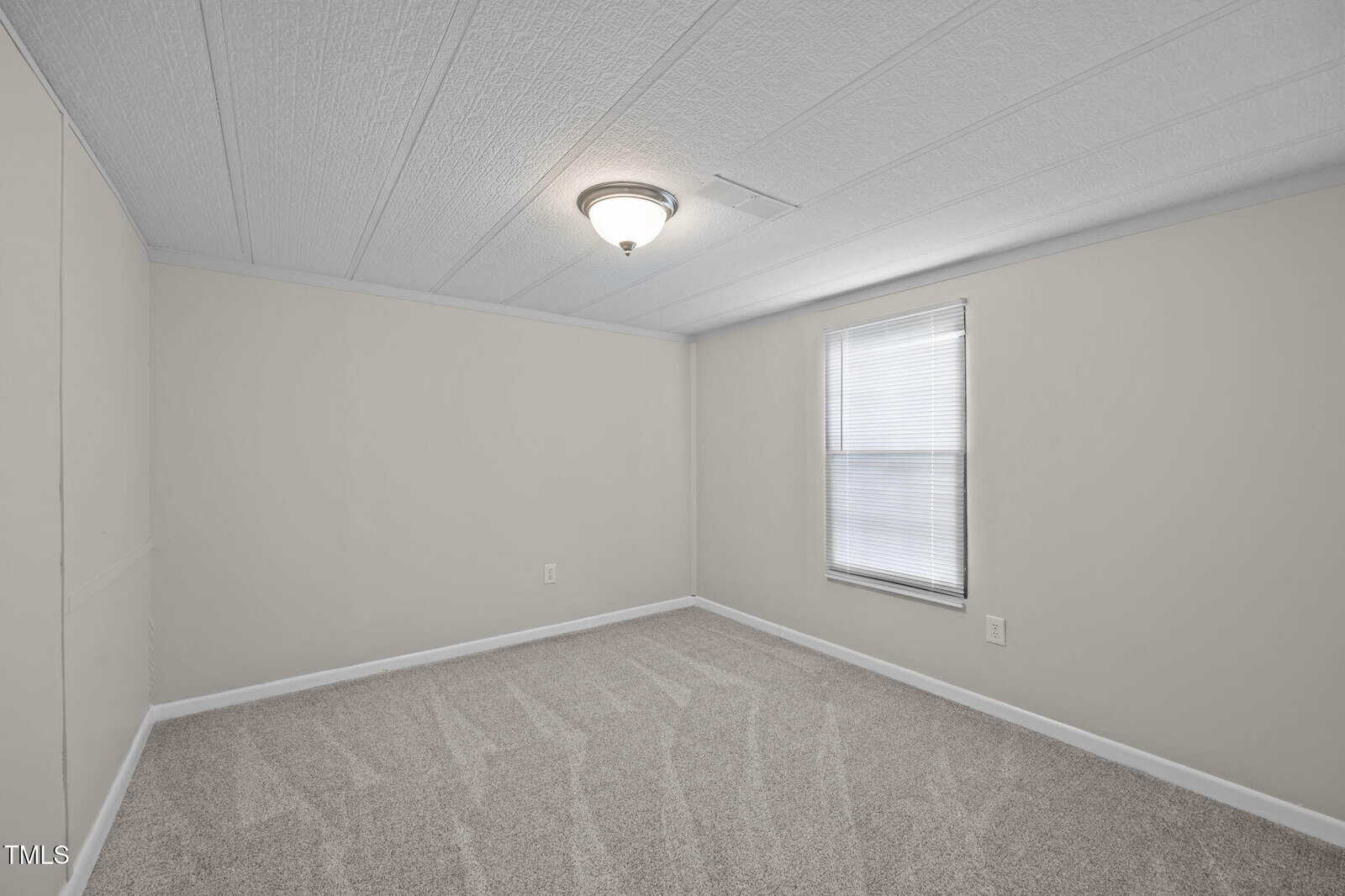 Empty room with light gray carpet, beige walls, a single window with closed blinds, and a ceiling-mounted light fixture. The room appears clean and well-lit.
