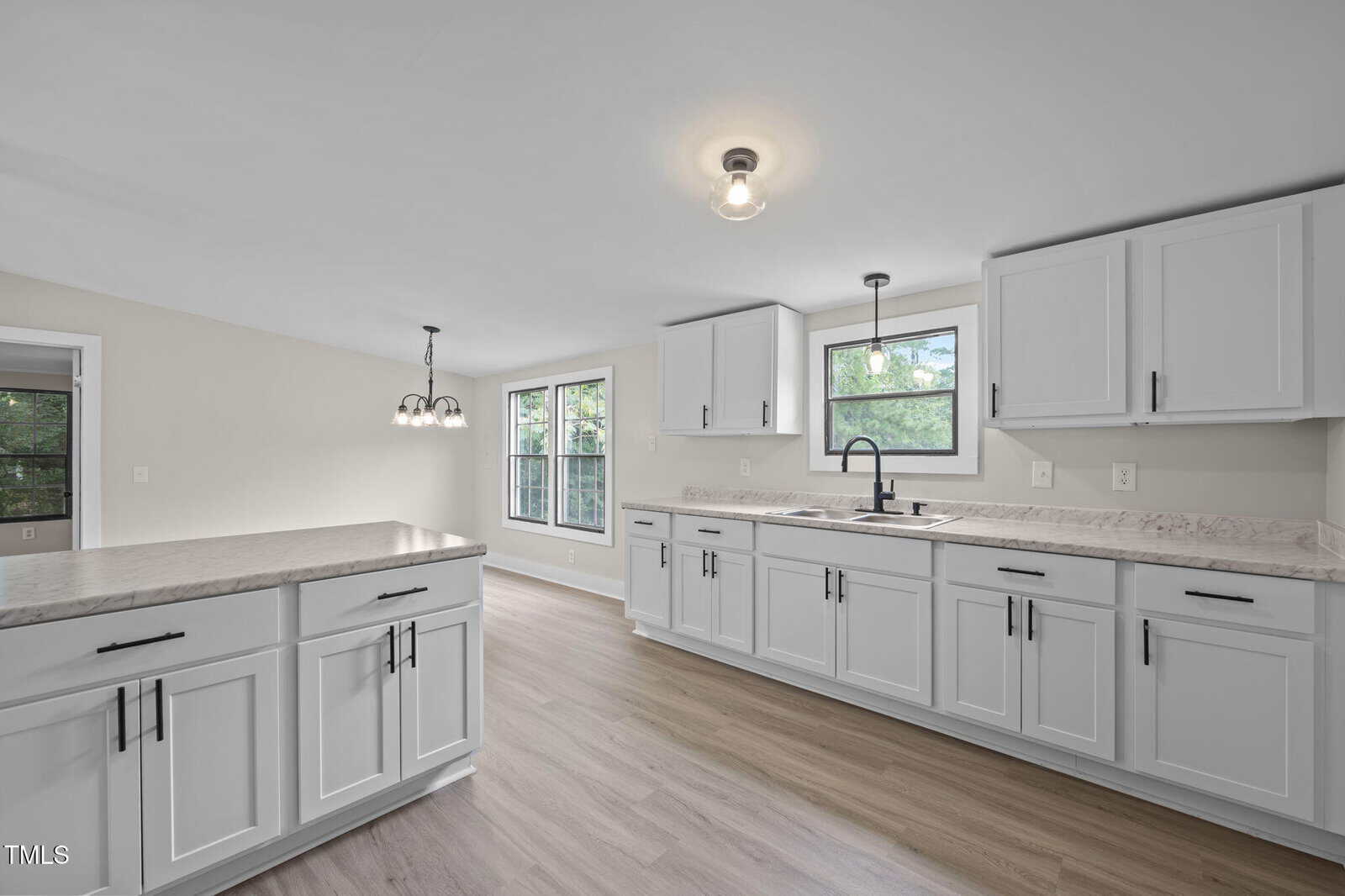 A modern kitchen with white cabinets, light-colored countertops, stainless steel fixtures, light wood flooring, and windows providing natural light. A hanging light fixture is above the adjoining dining area.