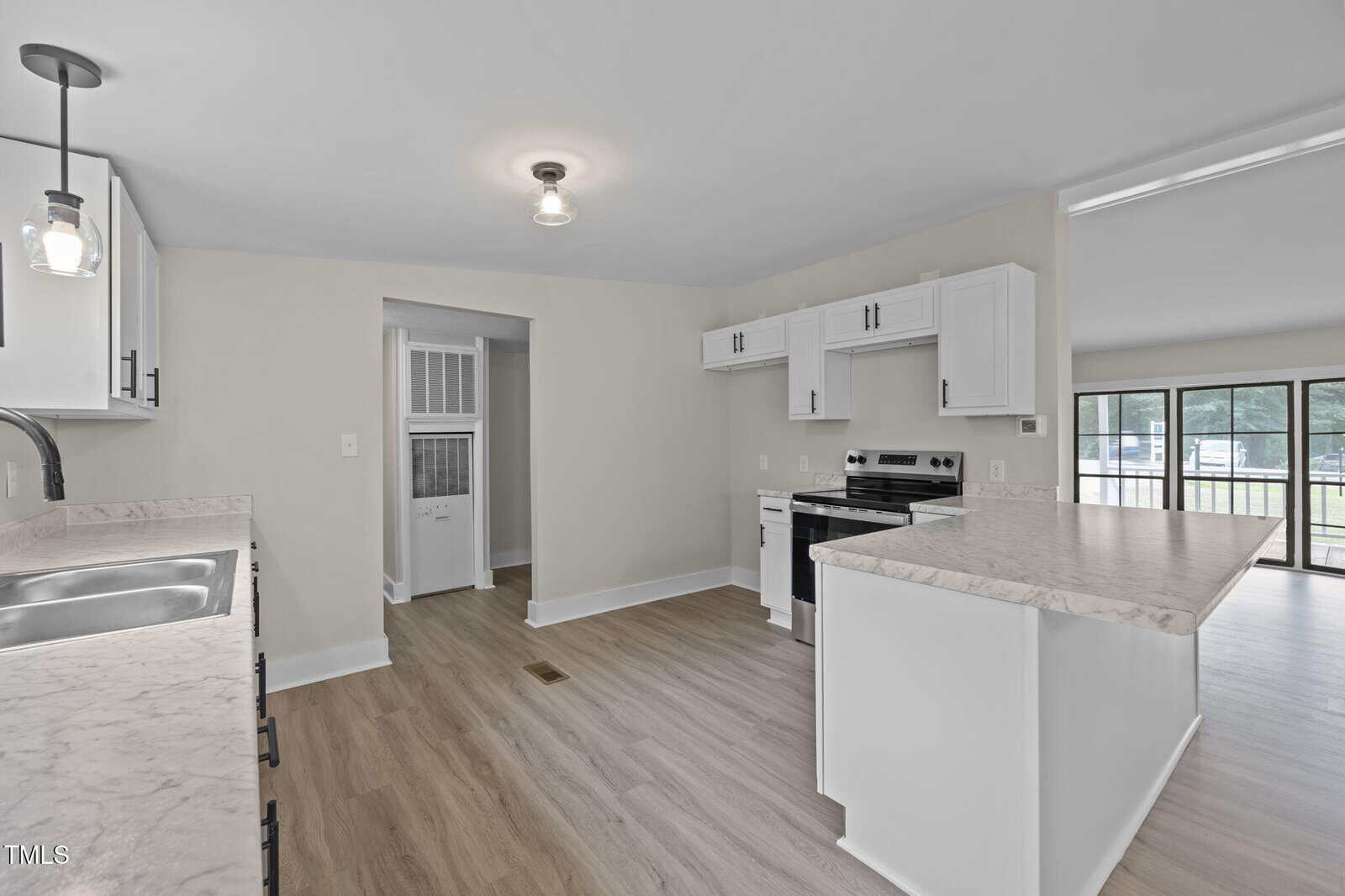 A modern kitchen with white cabinets, marble-look countertops, stainless steel appliances, and light wood flooring. An open doorway leads to another room, and large windows are visible in the background.