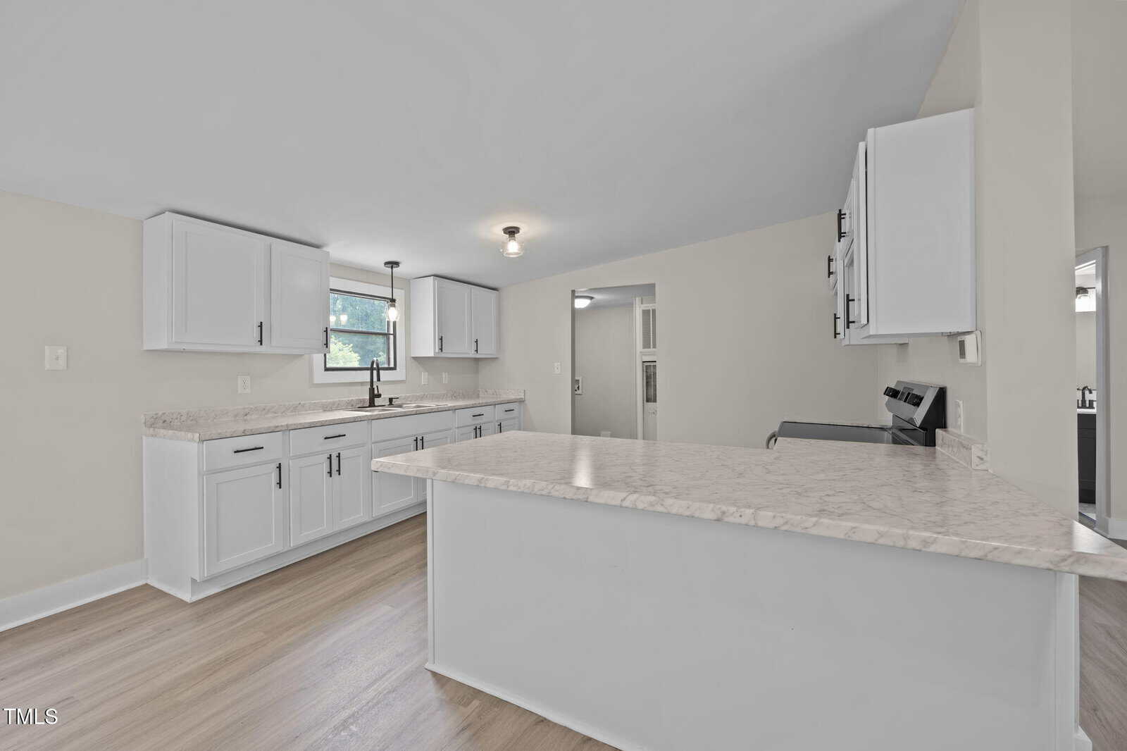A kitchen with white cabinets, light-colored countertops, stainless steel appliances, a window above the sink, and light wood flooring. The room is well-lit with ceiling lights and natural light.