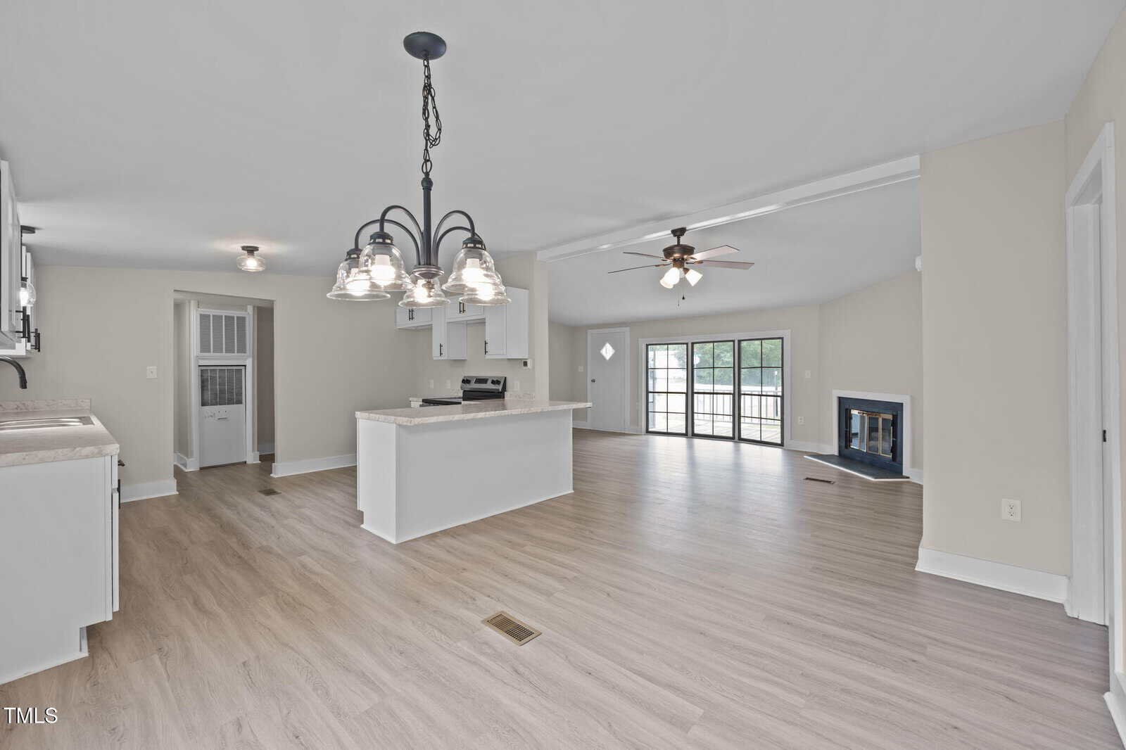 Spacious open-concept kitchen and living room with light wood floors, white cabinets, pendant lighting over an island, a ceiling fan, a modern fireplace, and large glass doors leading outside.