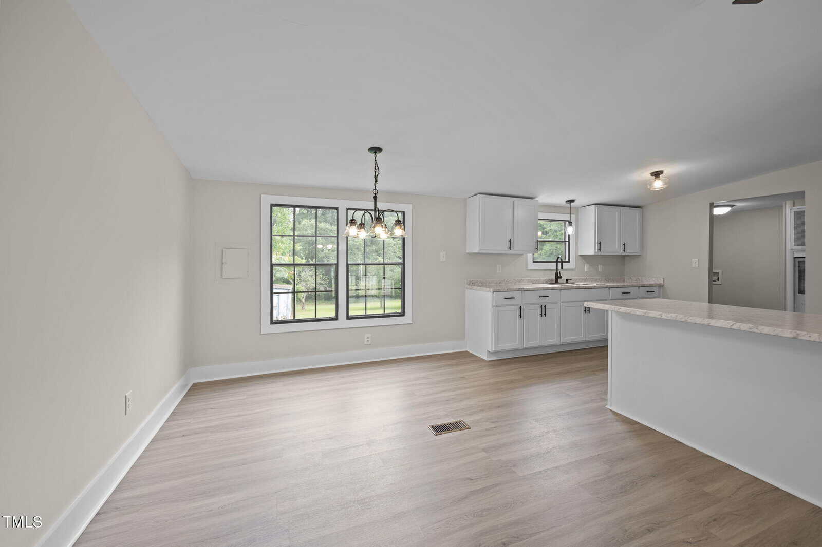 Open-concept kitchen and dining area with light wood flooring, white walls, and white cabinetry. Large window and glass pendant light in dining space; kitchen has a window above the sink and marble-look countertops.