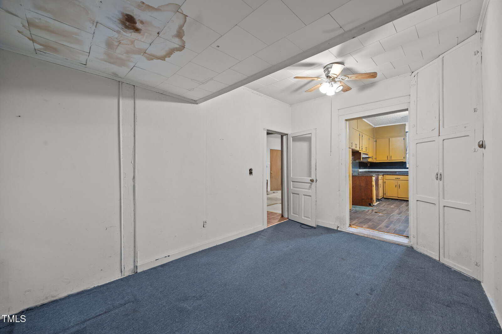 A room with blue carpet, white walls, a stained ceiling, built-in cabinets, and a ceiling fan, leading to a hallway and a kitchen with yellow cabinets and dark countertops.