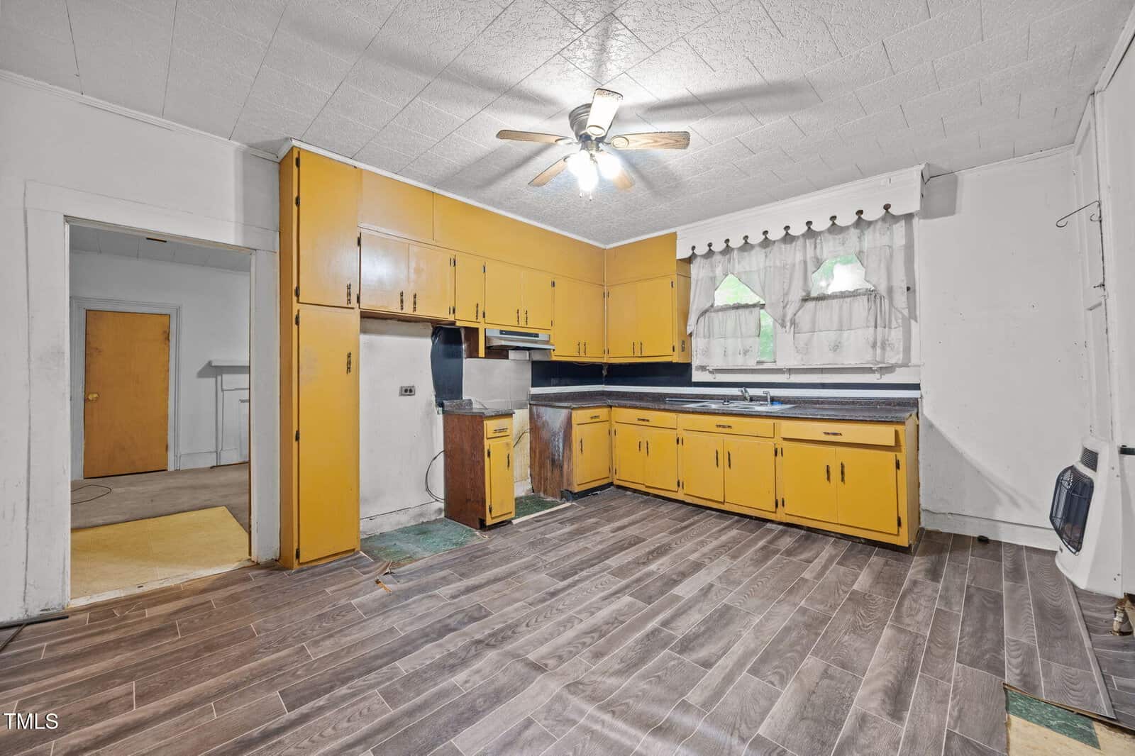 A kitchen with yellow cabinets, a dark countertop, wood-style flooring, a ceiling fan with a light, and two small windows with white curtains above the sink. The space appears worn and somewhat outdated.