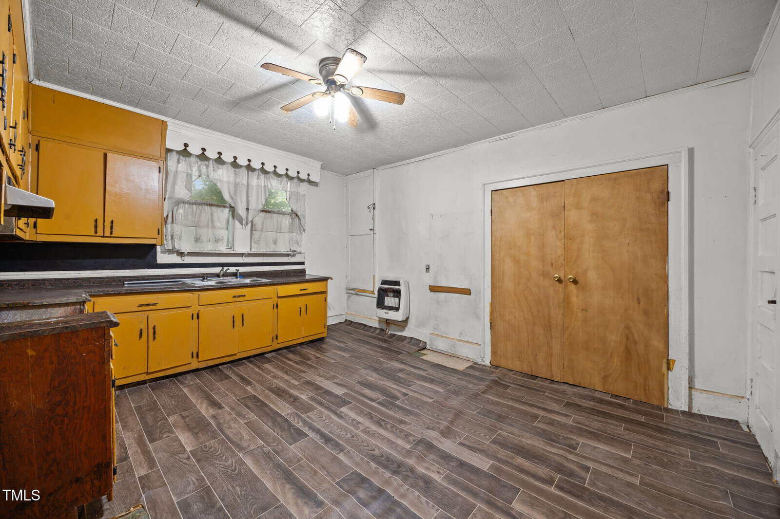 A kitchen with yellow cabinets, a dark countertop, wooden double doors, brown wood-look flooring, a ceiling fan with lights, and a window with sheer curtains and valance. The walls are white and show signs of wear.