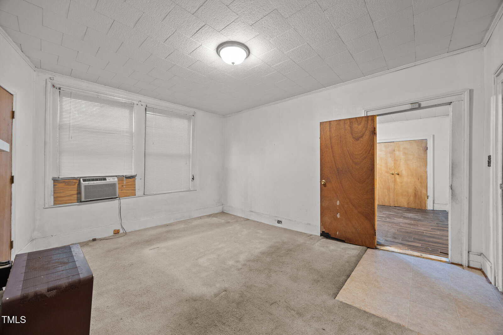 A mostly empty room with beige carpet, white walls, a ceiling light, window with blinds and an air conditioner, and an open wood door leading to a room with dark flooring.