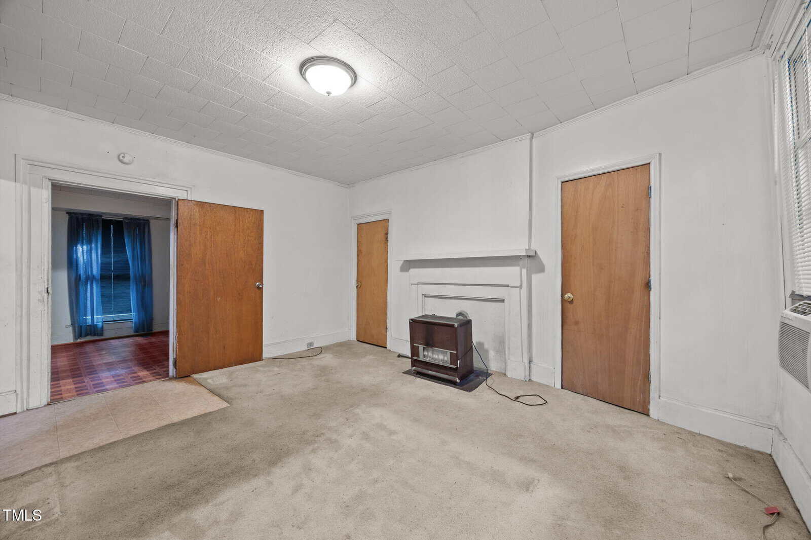 A mostly empty room with three wooden doors, beige carpet, white walls and ceiling, a wall heater under a mantel, and a window with blinds. An adjacent room with blue curtains is visible through an open doorway.