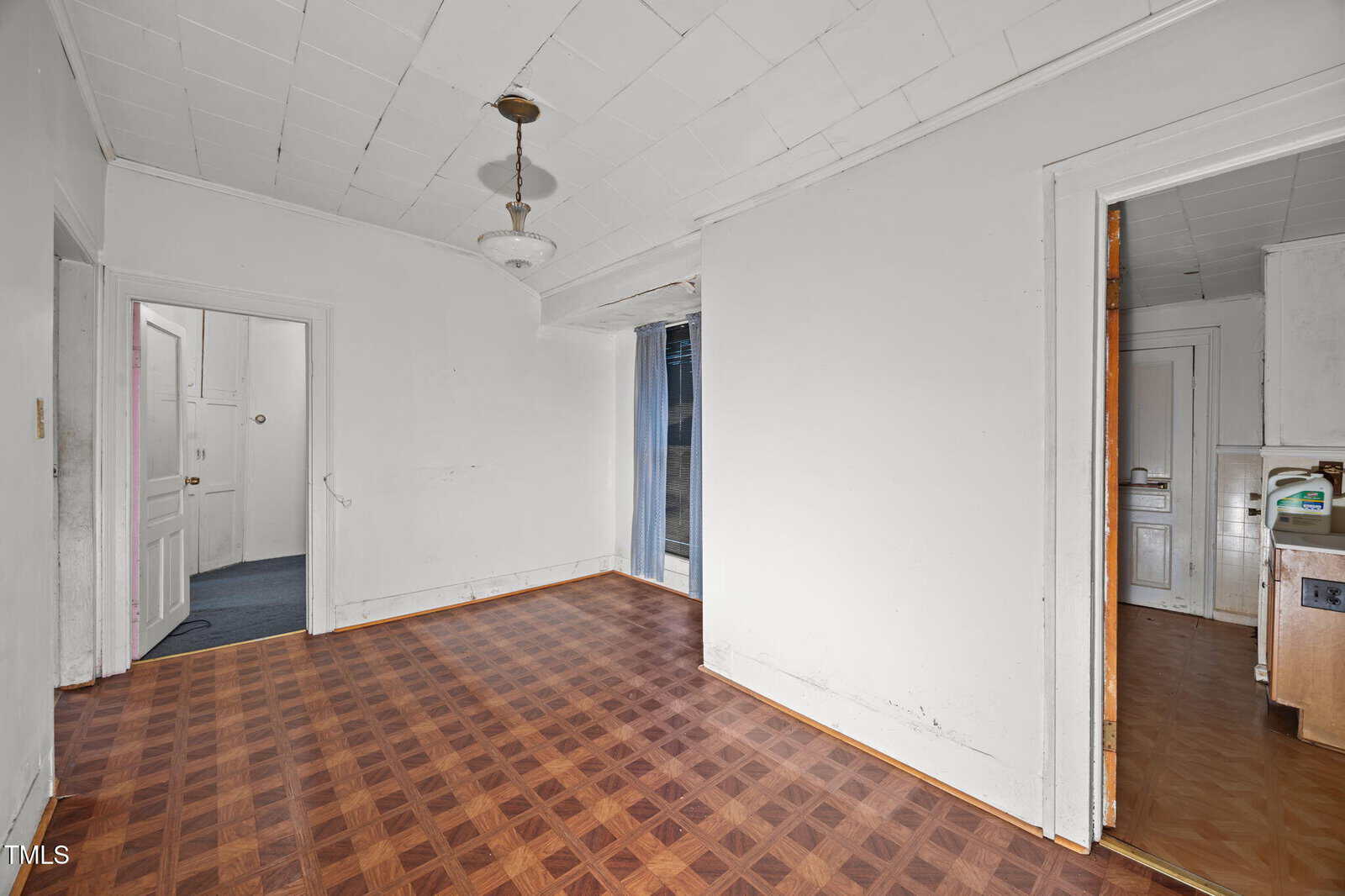 An empty room with white walls, a patterned wooden floor, a ceiling light fixture, and doorways leading to other rooms. One door opens to a kitchen, another to a hallway, and there’s a sliding glass door.