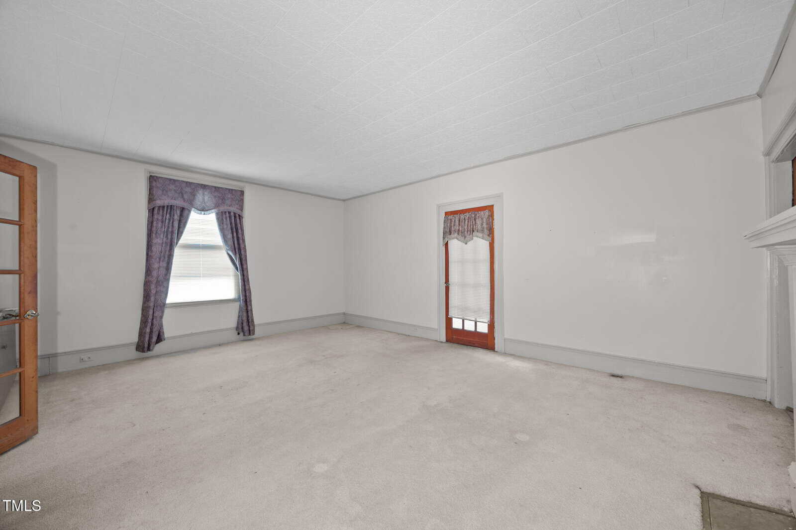 Empty room with light-colored carpet, white walls, and a white ceiling. There is a window with purple curtains, a wooden door with a matching curtain, a glass-paned door, and a fireplace on the right side.