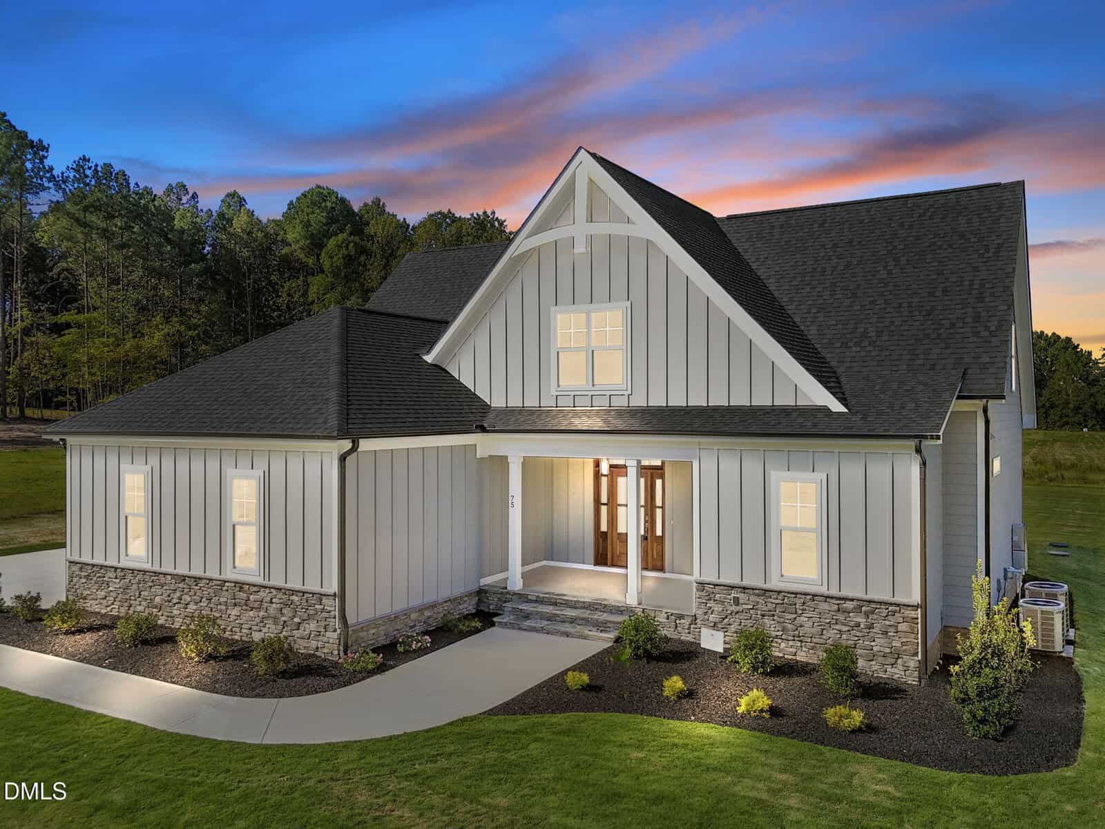 A modern two-story house at 75 Broadleaf Lane, Youngsville, features gray vertical siding, stone accents, and a dark shingle roof at sunset. The entryway glows as the driveway curves before a landscaped lawn bordered by trees.