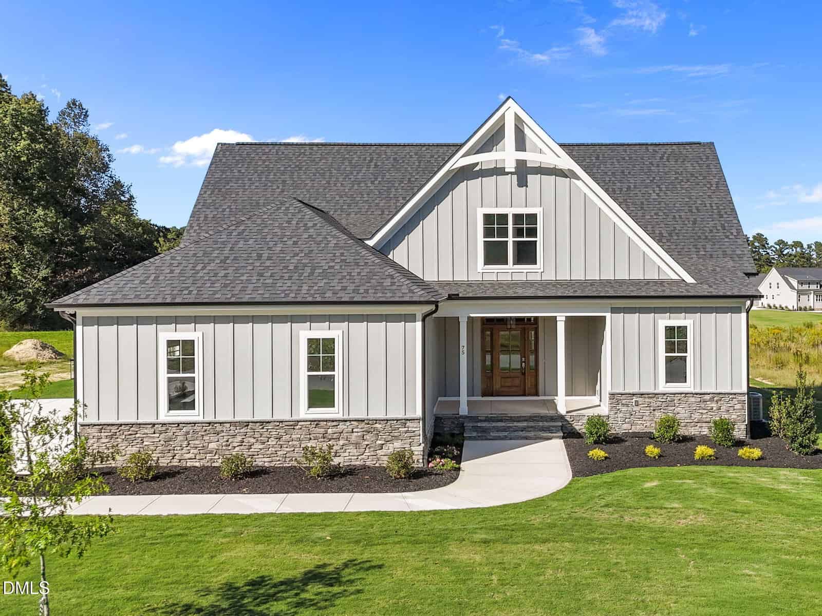 A modern two-story house with light gray siding, stone accents on the lower exterior, a dark shingled roof, white trim, a covered front porch, and neatly landscaped lawn under a clear blue sky.