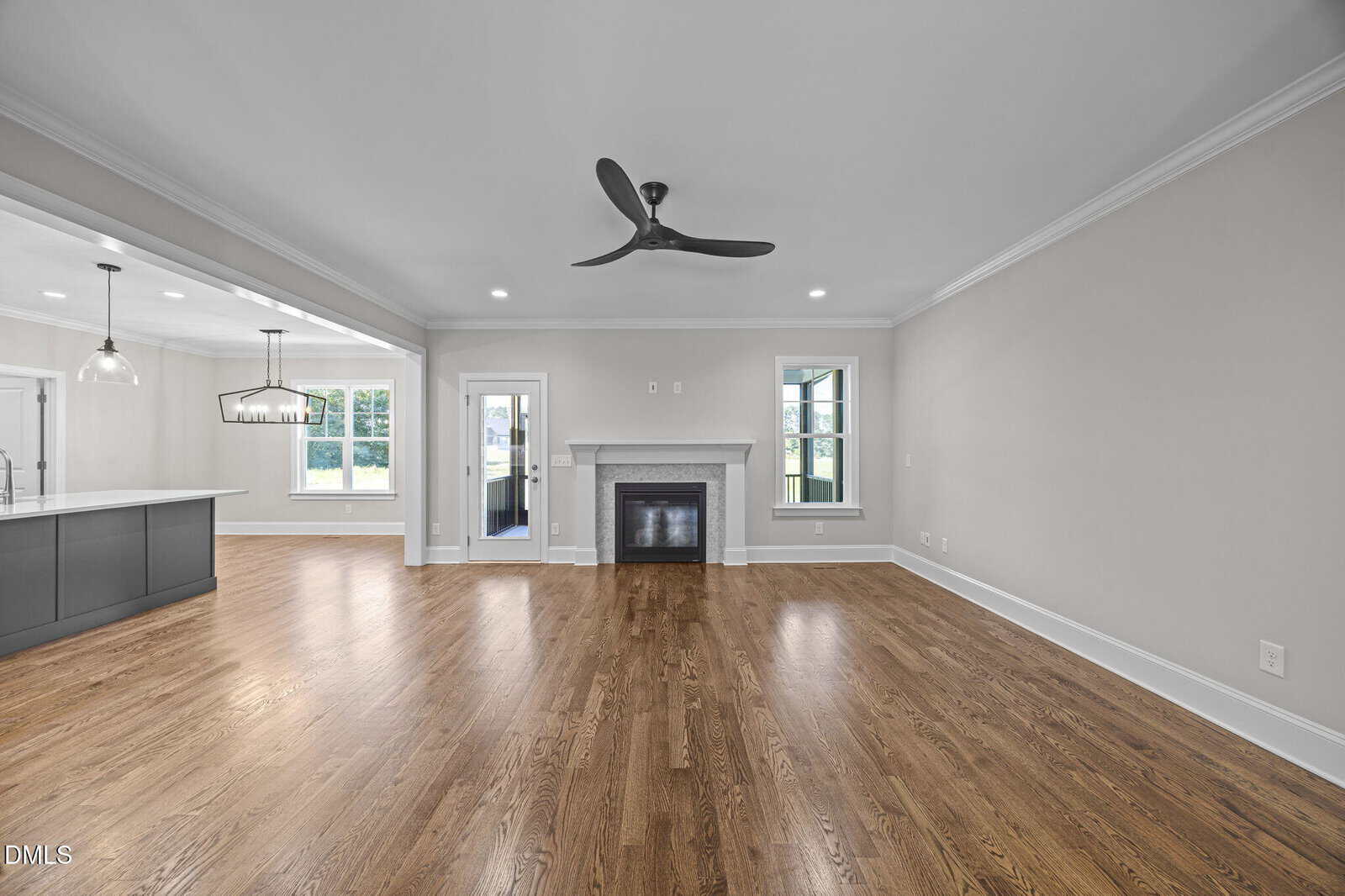 Spacious living area with hardwood floors, a ceiling fan, a fireplace, large windows, and an open kitchen with pendant lighting and an island.