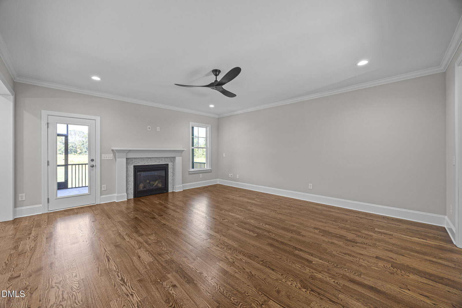Spacious empty living room with hardwood floors, light gray walls, a black ceiling fan, white trim, a fireplace, and two windows. A door leads to an outdoor area with a view of trees.