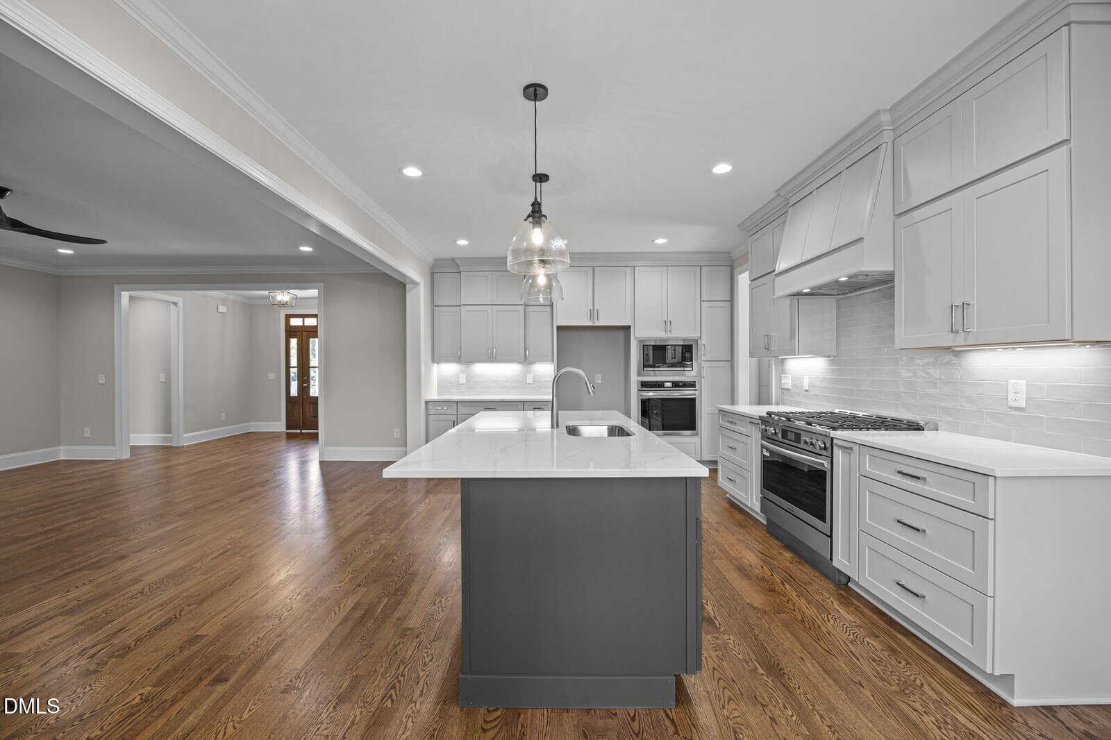 Modern kitchen with white cabinets, stainless steel appliances, large center island with sink, pendant lights, hardwood floors, and an open floor plan leading to another room and an entryway in the background.