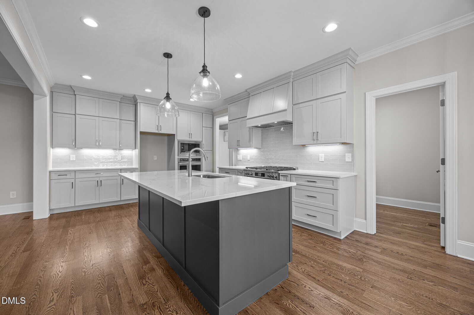 A modern kitchen with white cabinets, a gray island, stainless steel appliances, pendant lights, white countertops, and wooden floors. The room is well-lit with recessed ceiling lights and natural light.