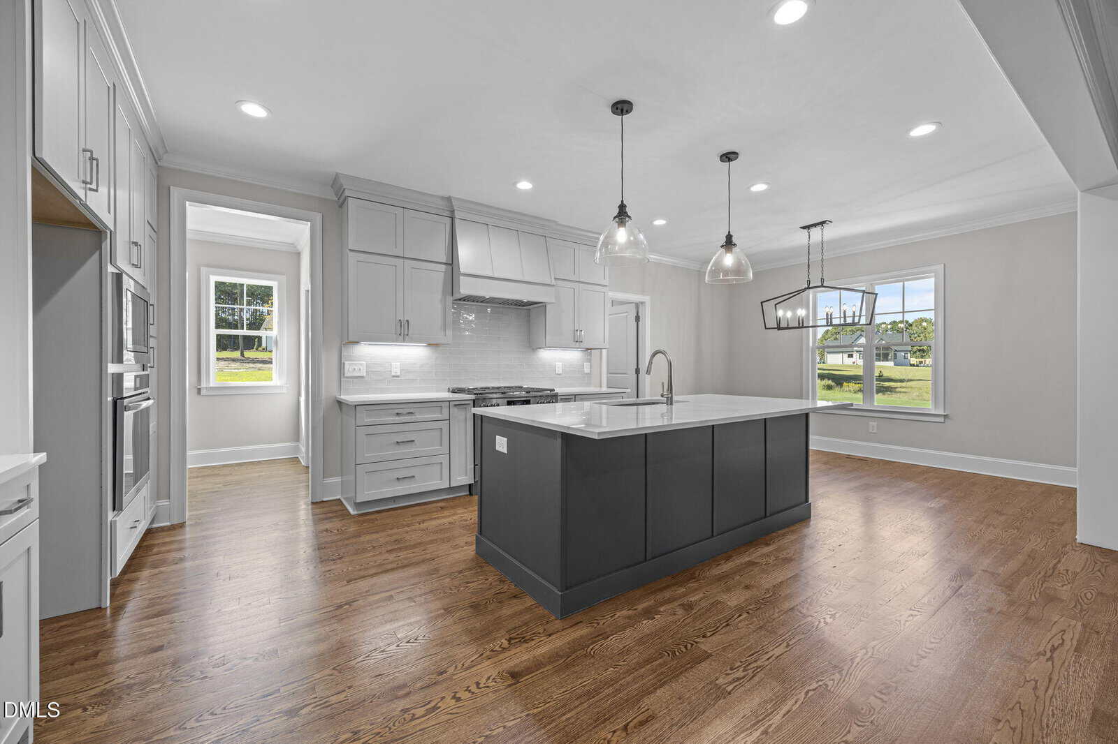 Modern kitchen with light gray cabinets, white subway tile backsplash, a dark gray island with a white countertop, stainless steel appliances, pendant lights, wood flooring, and large windows providing natural light.