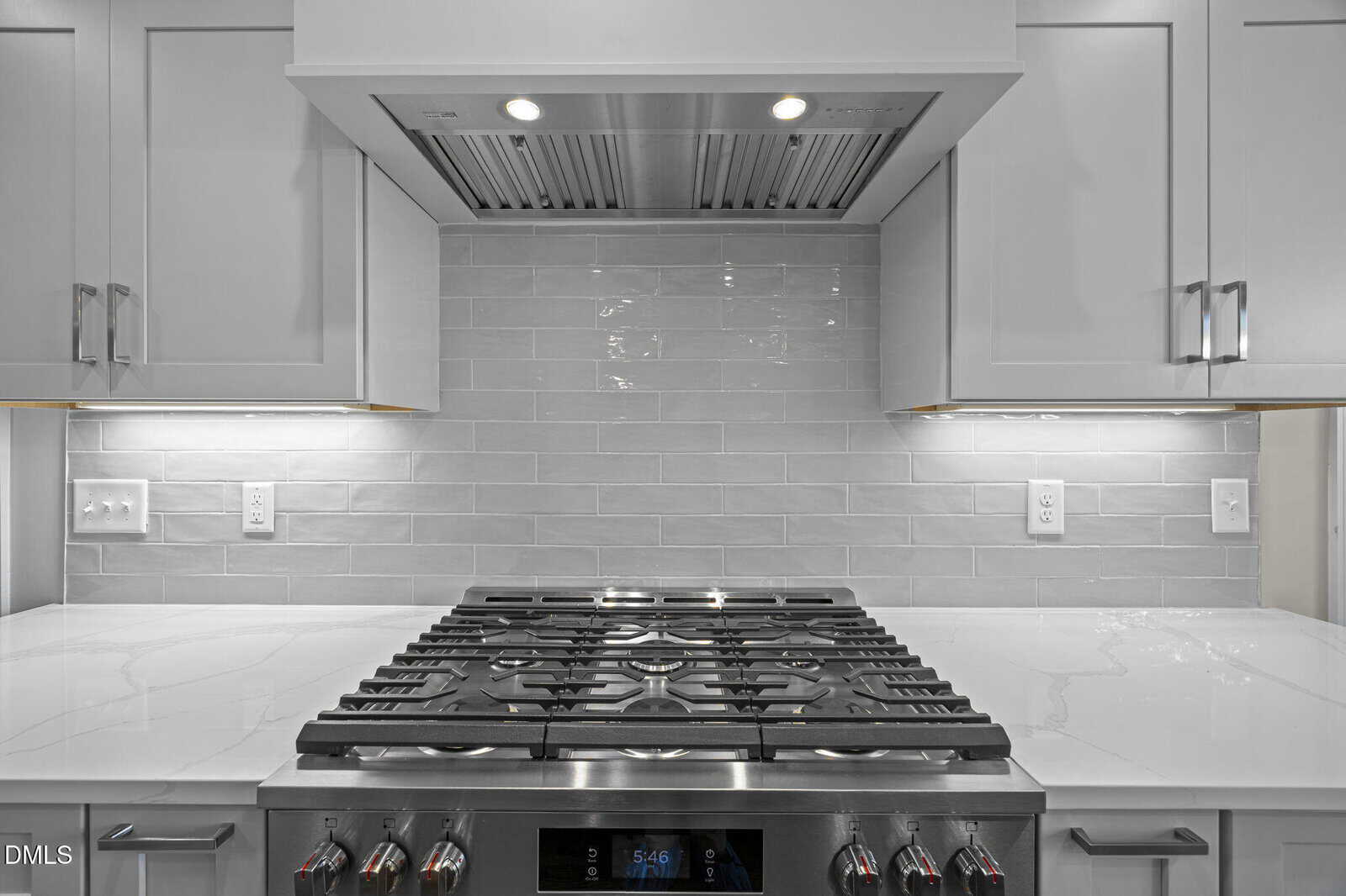 A stainless steel gas cooktop with red-control knobs is set into a white countertop beneath a range hood. The backsplash features light gray subway tiles, and white upper cabinets are installed above.