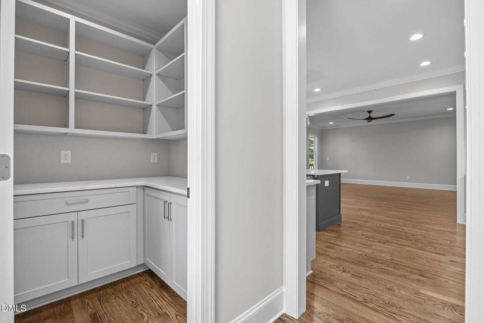 A white walk-in pantry with open shelving and cabinets is shown next to a hallway that leads to a spacious living area with hardwood floors and neutral-colored walls.